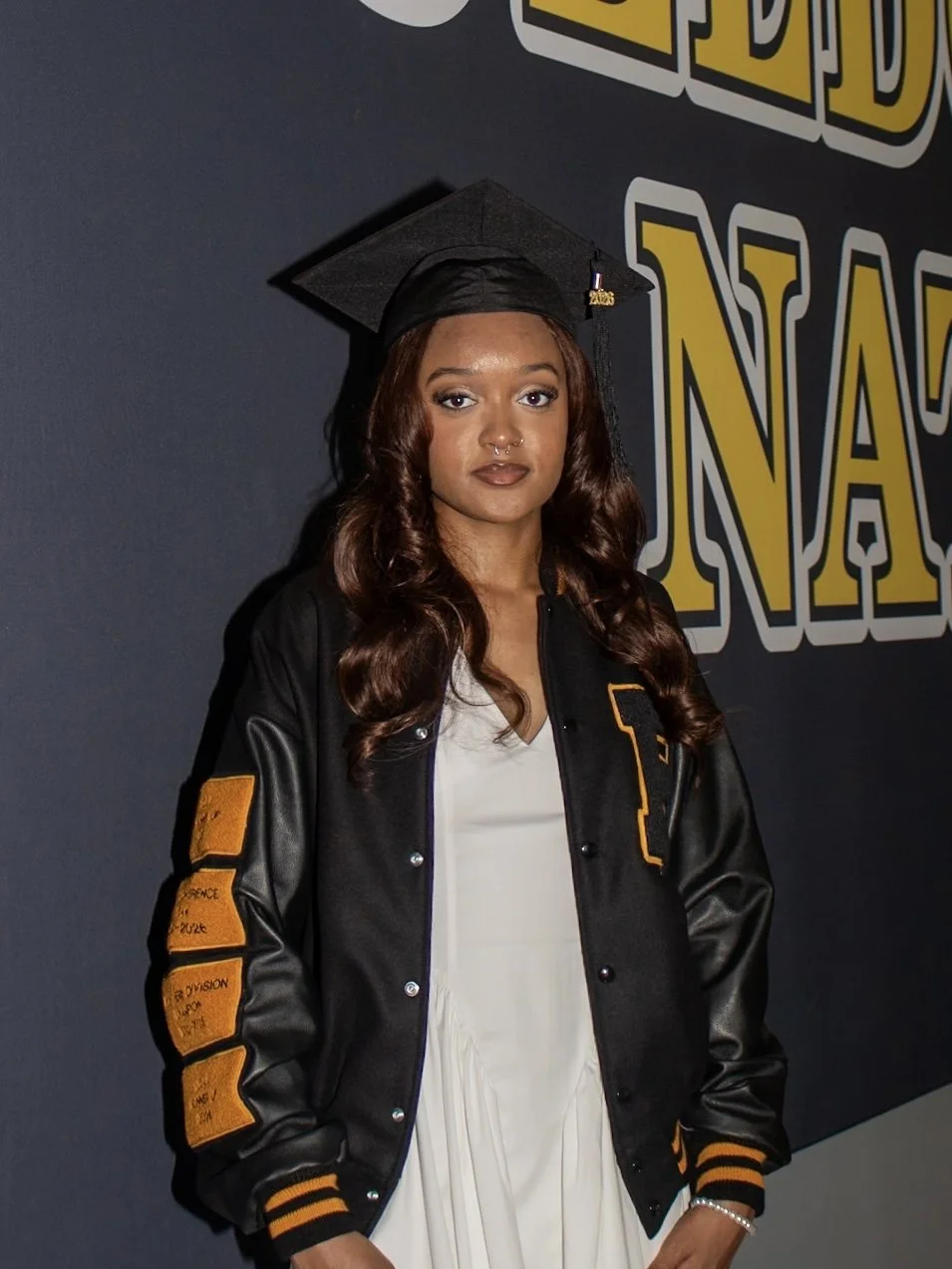 A young woman in a graduation cap and gown, wearing a letterman jacket, standing in front of a wall with large yellow and white text that says "Graduation".