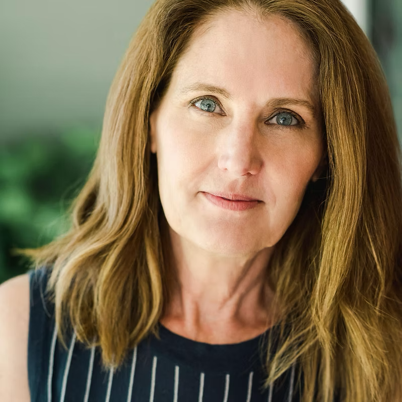 Close-up portrait of a woman with long, wavy red hair and blue eyes, wearing a navy sleeveless top with white stripes, outdoors with blurred green background.