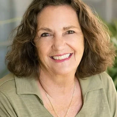 A smiling woman with shoulder-length curly brown hair, wearing a light beige top and a gold necklace, in an indoor setting with blurred background.