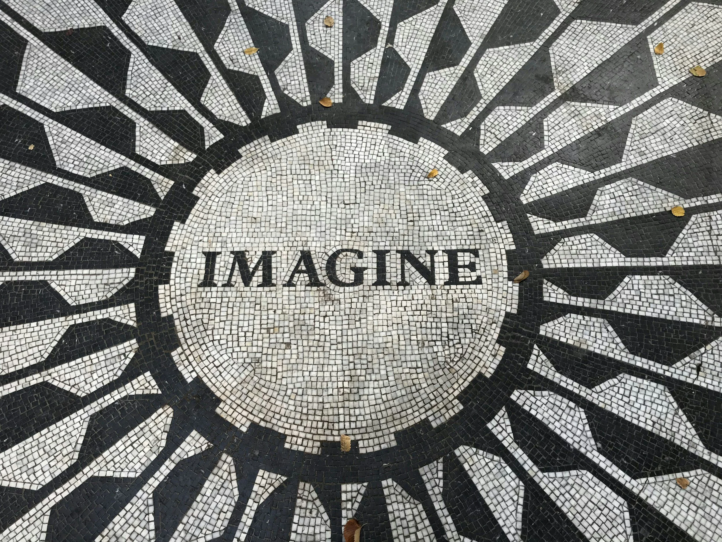 Close-up of black and white mosaic flooring with a radial pattern and the word 'IMAGINE' embedded in the design, with some small fallen leaves on the surface.