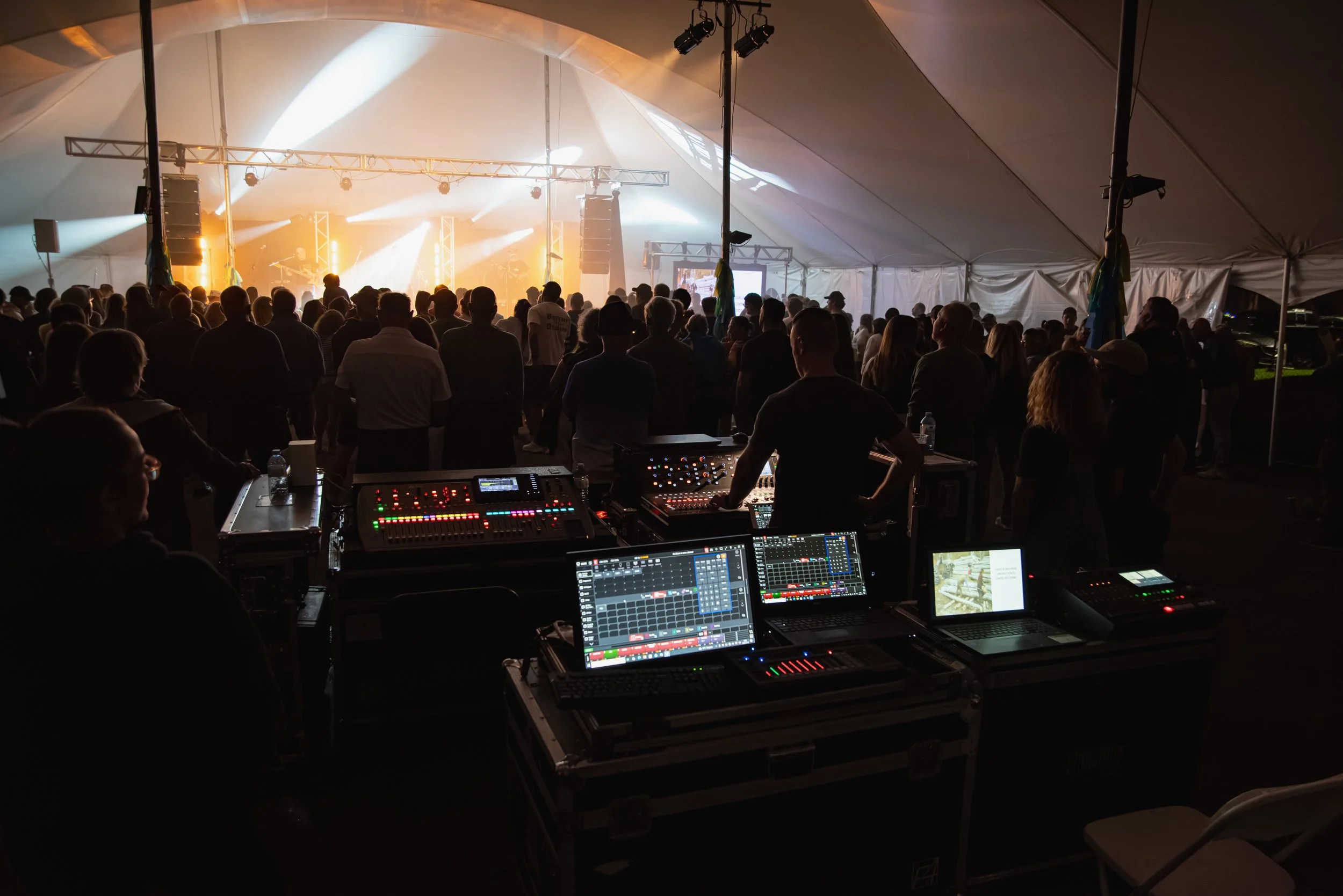 Une grande tente blanche remplie de personnes regardant un concert ou un événement avec un éclairage coloré. À l'avant, il y a une console de mixage et du matériel audio avec des équipements de son et des écrans.