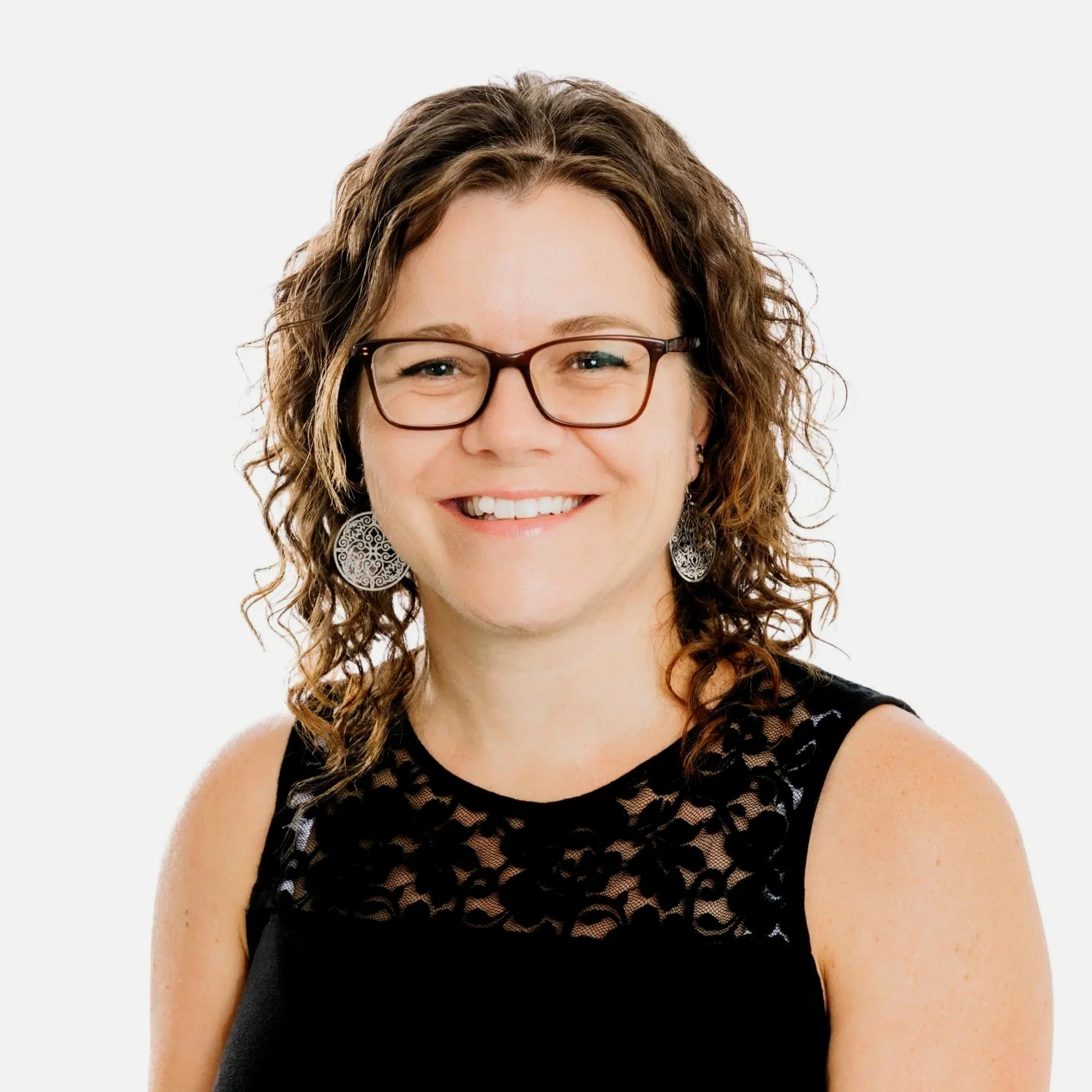 Portrait d'une femme souriante portant des lunettes, avec des boucles de cheveux bruns et une robe noire en dentelle, contre un fond blanc.