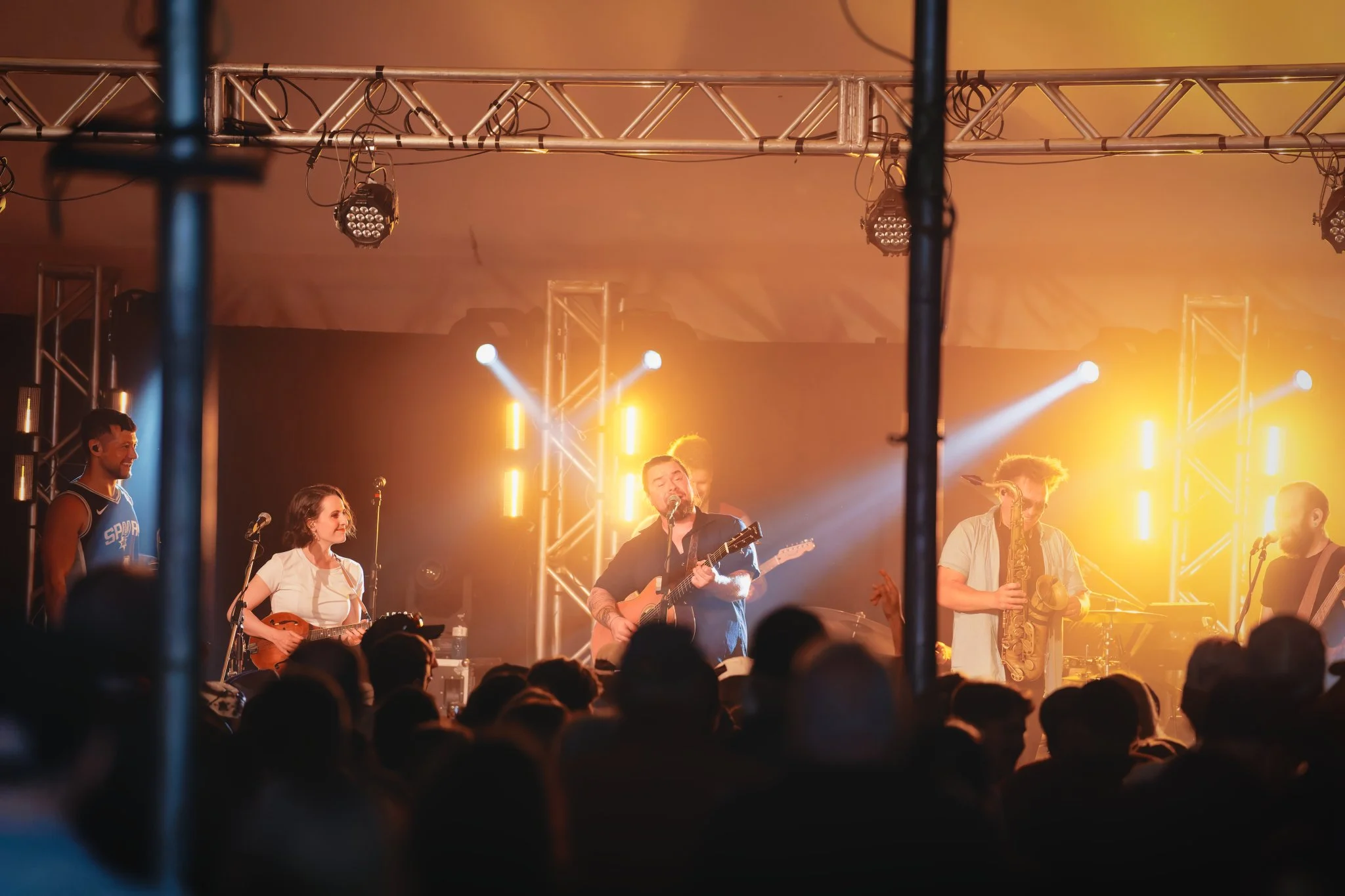 Un groupe de musiciens joue en concert sur scène, éclairée par des lumières jaunes et blanches, avec un public devant eux.
