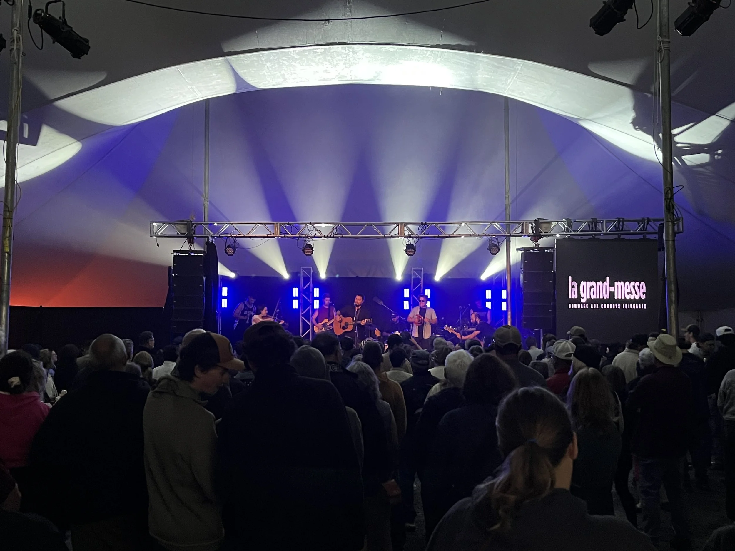Une scène de concert à l'intérieur avec des musiciens jouant sur scène, une audience devant eux, et un éclairage de scène coloré.