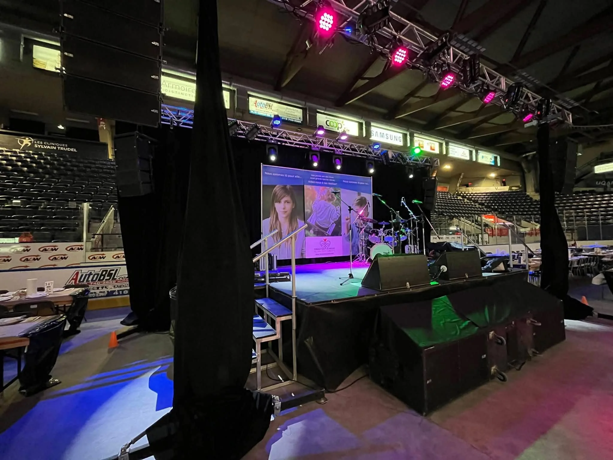 Une scène d'un événement ou concert dans une salle intérieure, avec une scène équipée de microphones et de spotlights colorés, des sièges vides, et des panneaux publicitaires en arrière-plan.