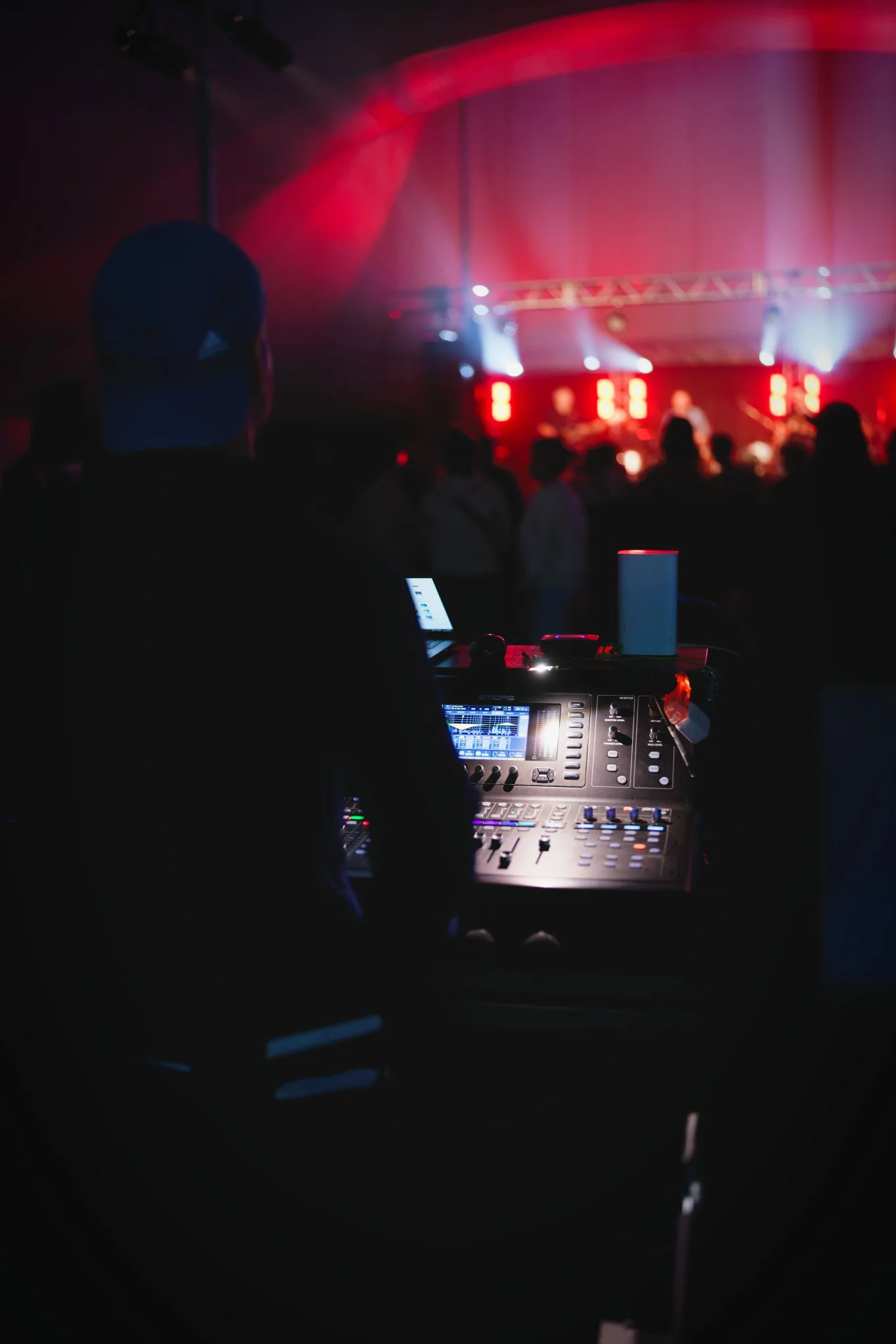Vue de la console de mixage audio lors d'une performance en direct, avec des lumières de scène en arrière-plan et des spectateurs silhouettes en premier plan.
