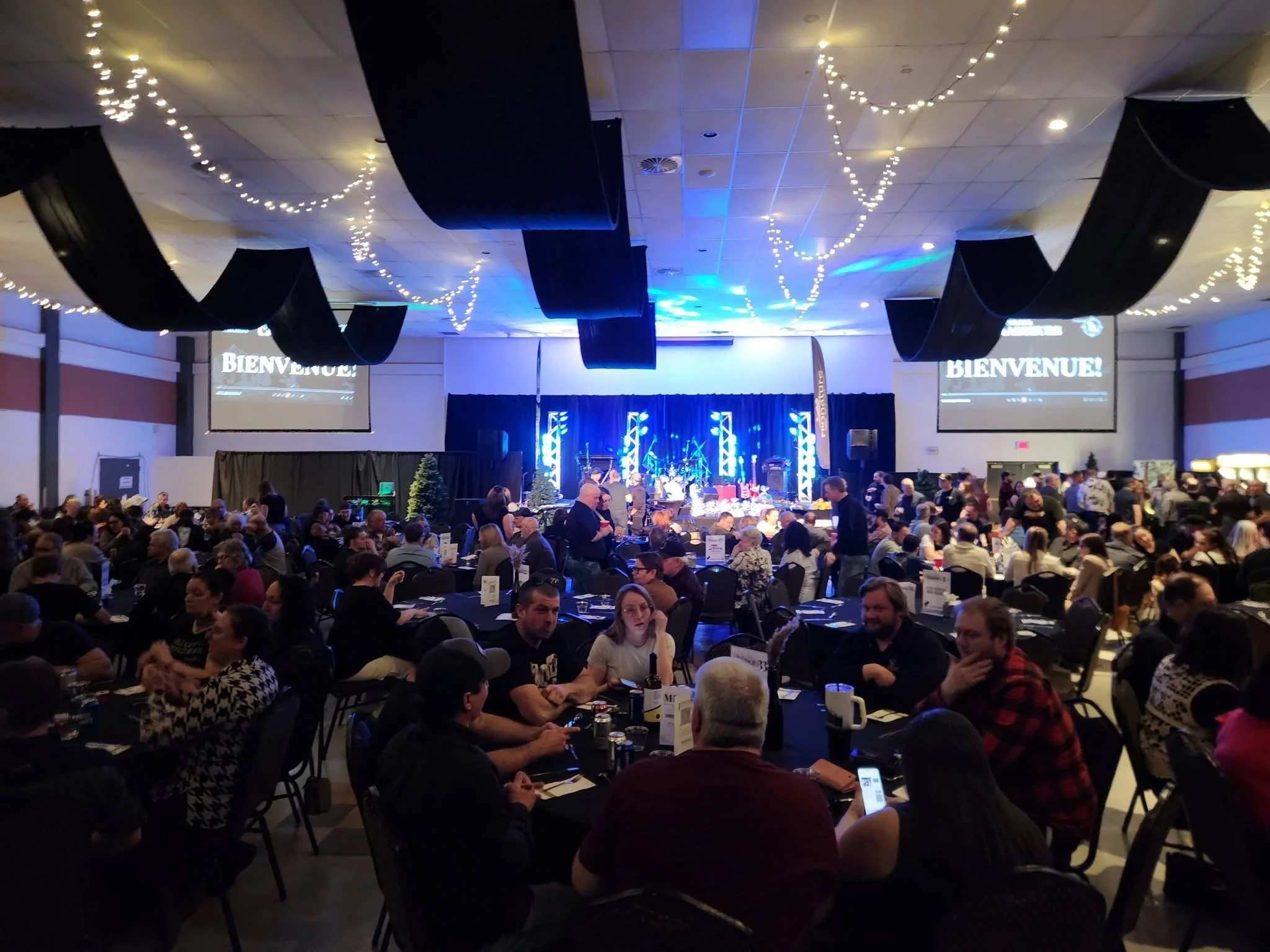 Une grande salle accueillant un nombre important de personnes assises autour de tables, avec un décor festif, des guirlandes lumineuses et une scène avec de la musique. Écran affichant le mot 'BIENVENUE !' à l'avant de la salle.