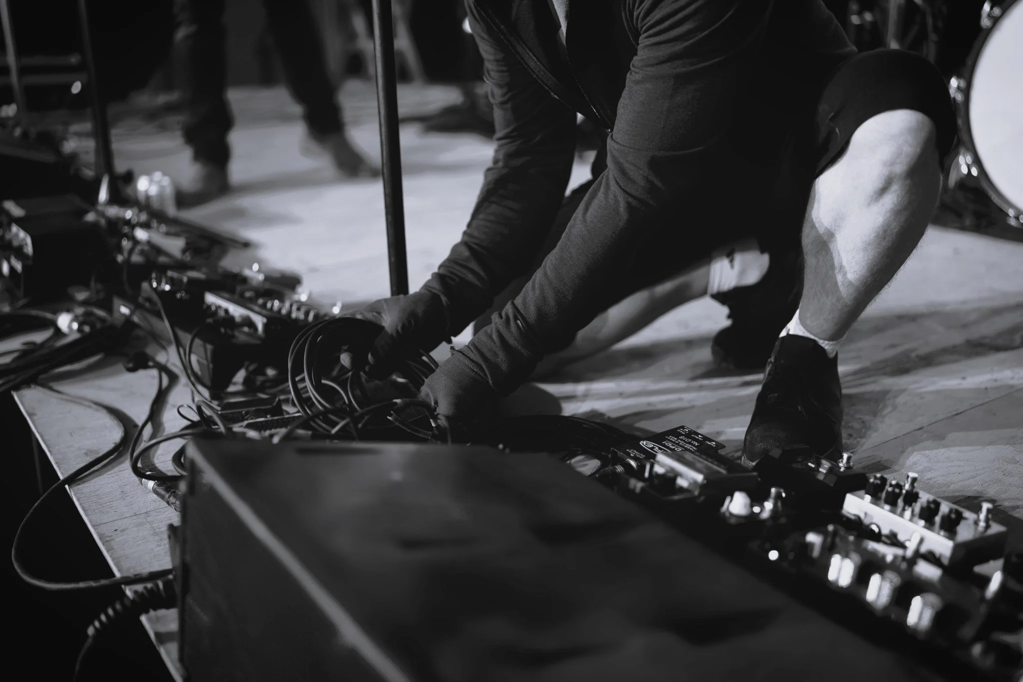 Une personne en pantalon sombre et bottes noires, occupée à manipuler du matériel électronique ou musical avec de nombreux câbles.