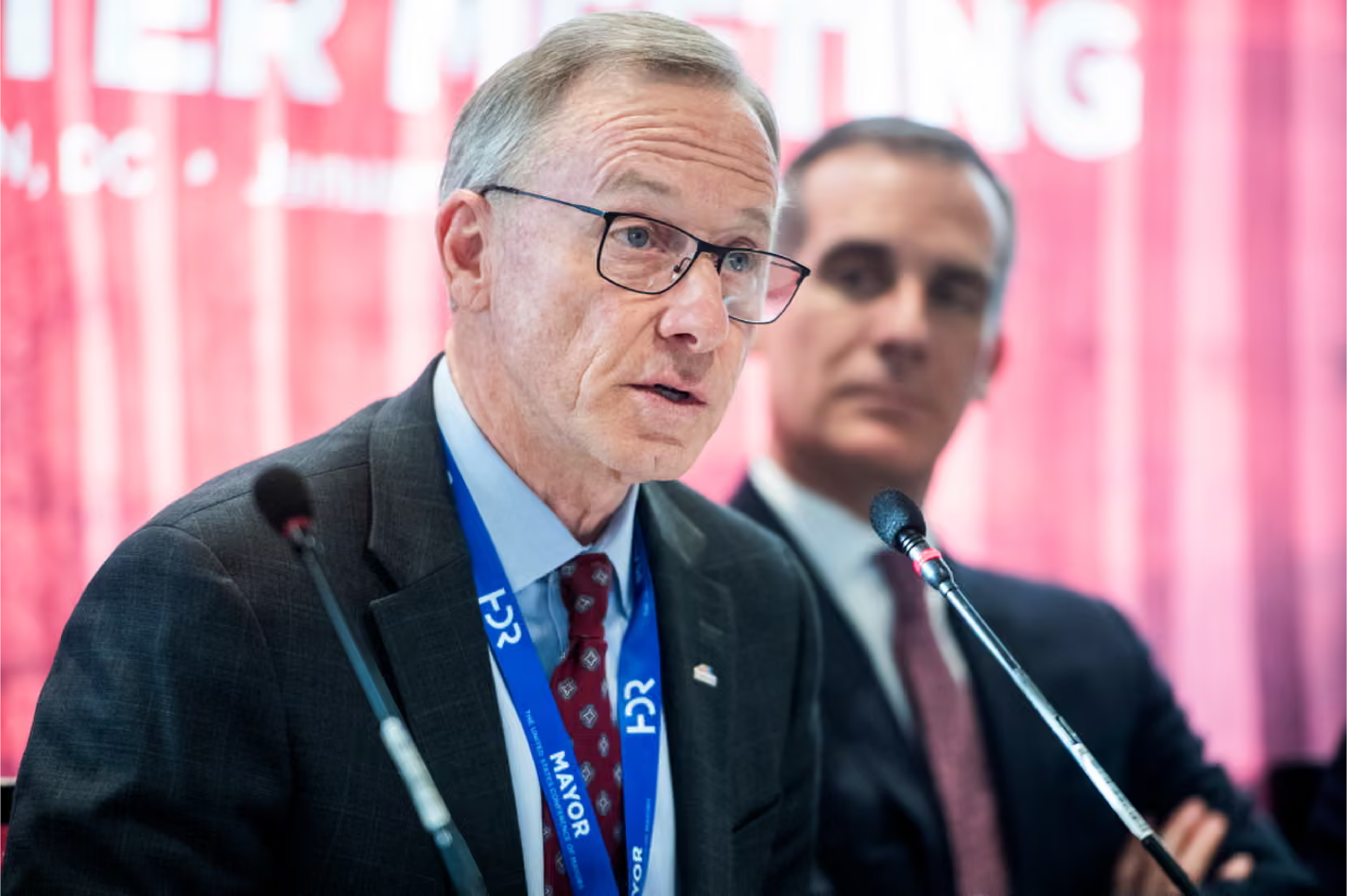 A man wearing glasses and a suit speaking at a microphone during a conference or political event, with another man blurred in the background, and pink banners in the background.