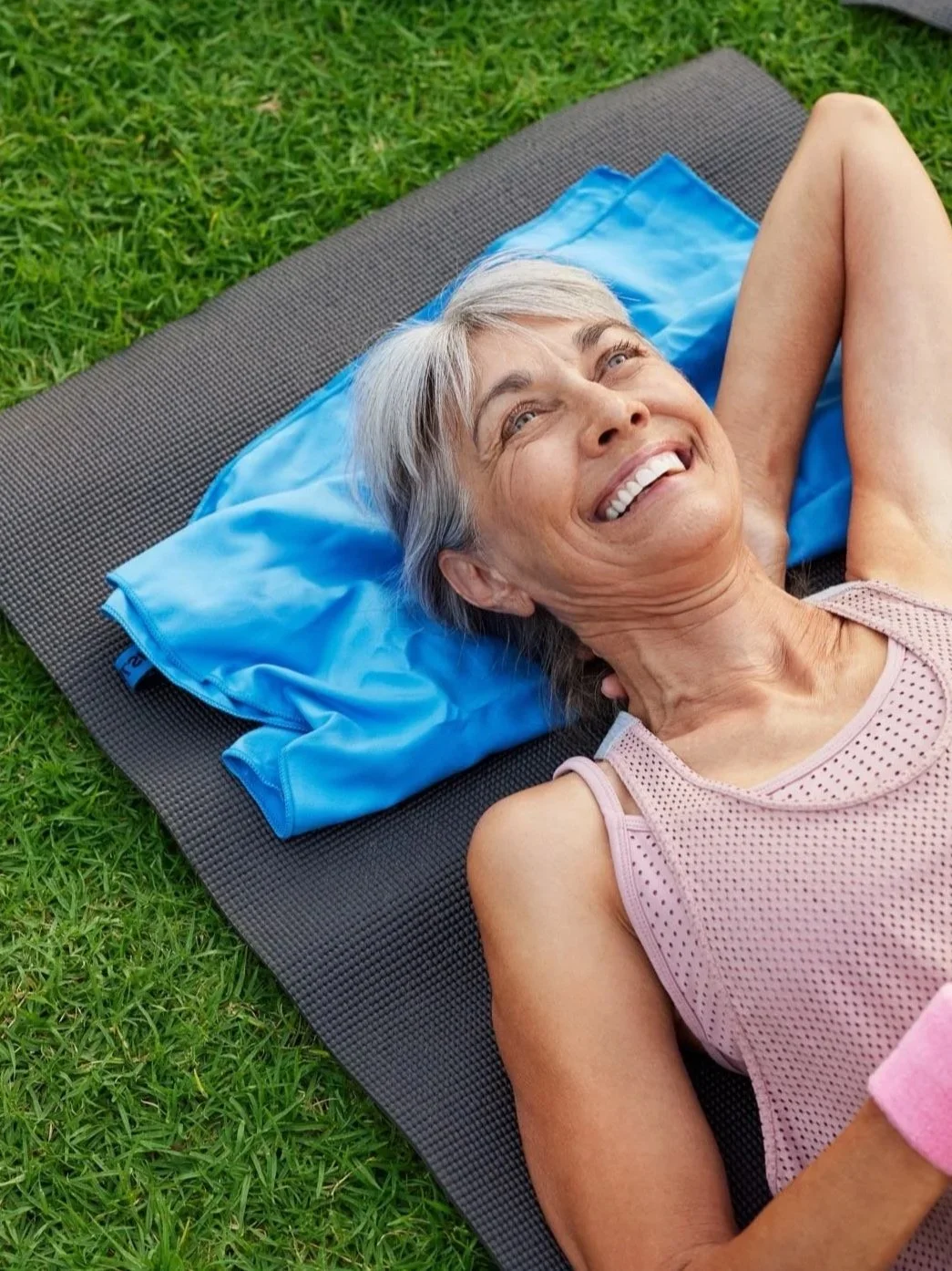 Smiling older woman lying on a black yoga mat on green grass, resting her head on a blue pillow, looking up with a relaxed and happy expression.