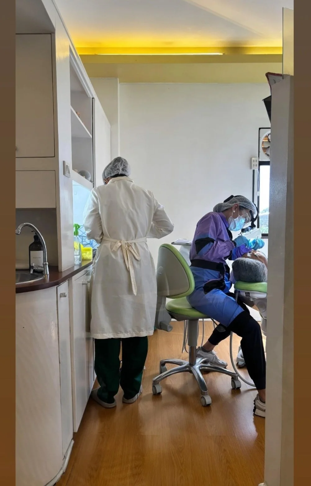 Medical professionals performing a dental procedure in a clinic, with one dentist seated at a patient chair and another standing nearby in a white coat, both wearing protective gear including masks and hair covers.