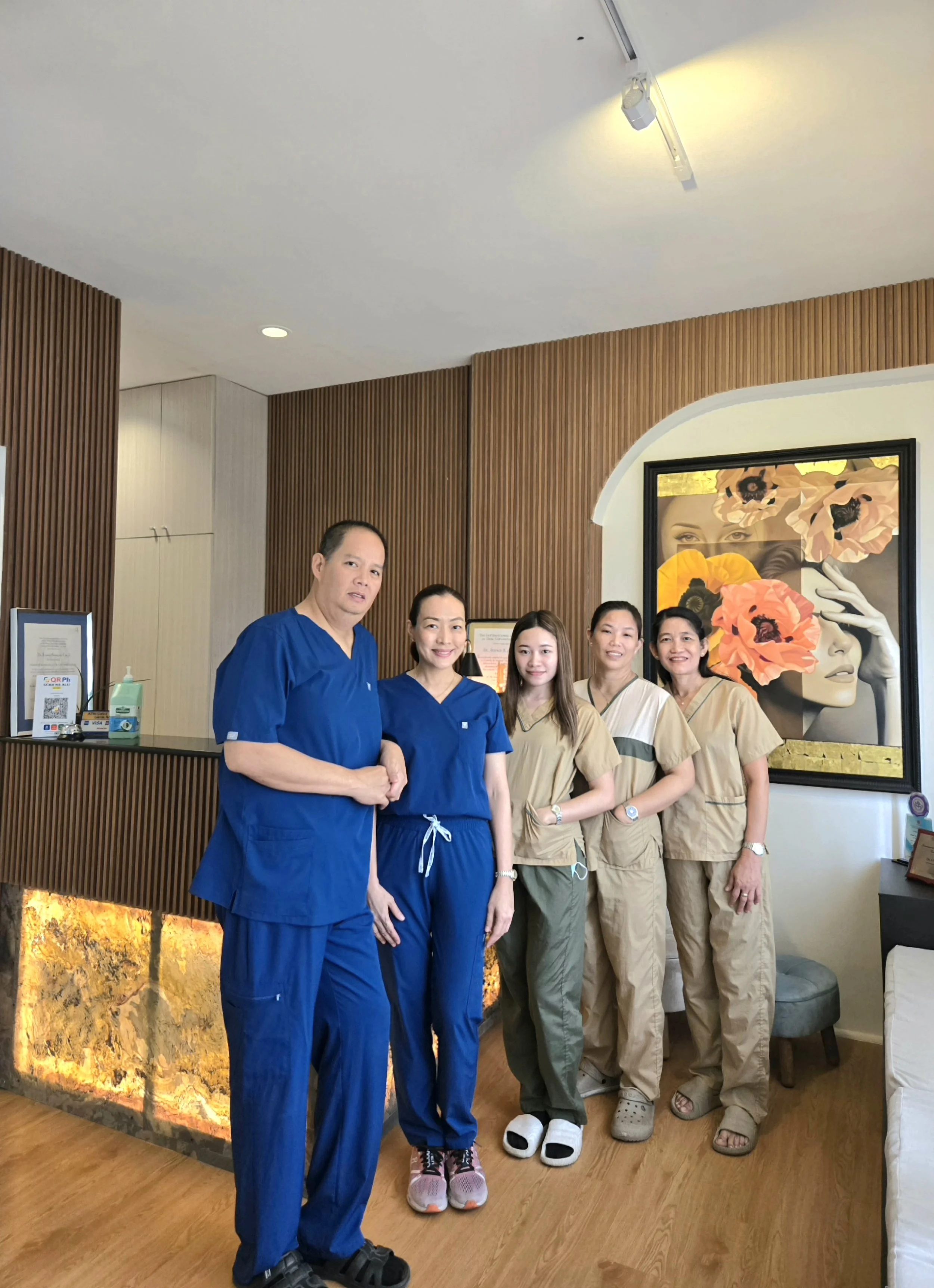 A group of six people, five women and one man, standing in a row inside a modern, warmly lit room with wooden wall accents and a large colorful floral painting. The group is dressed in casual and medical scrubs, smiling for the photo.