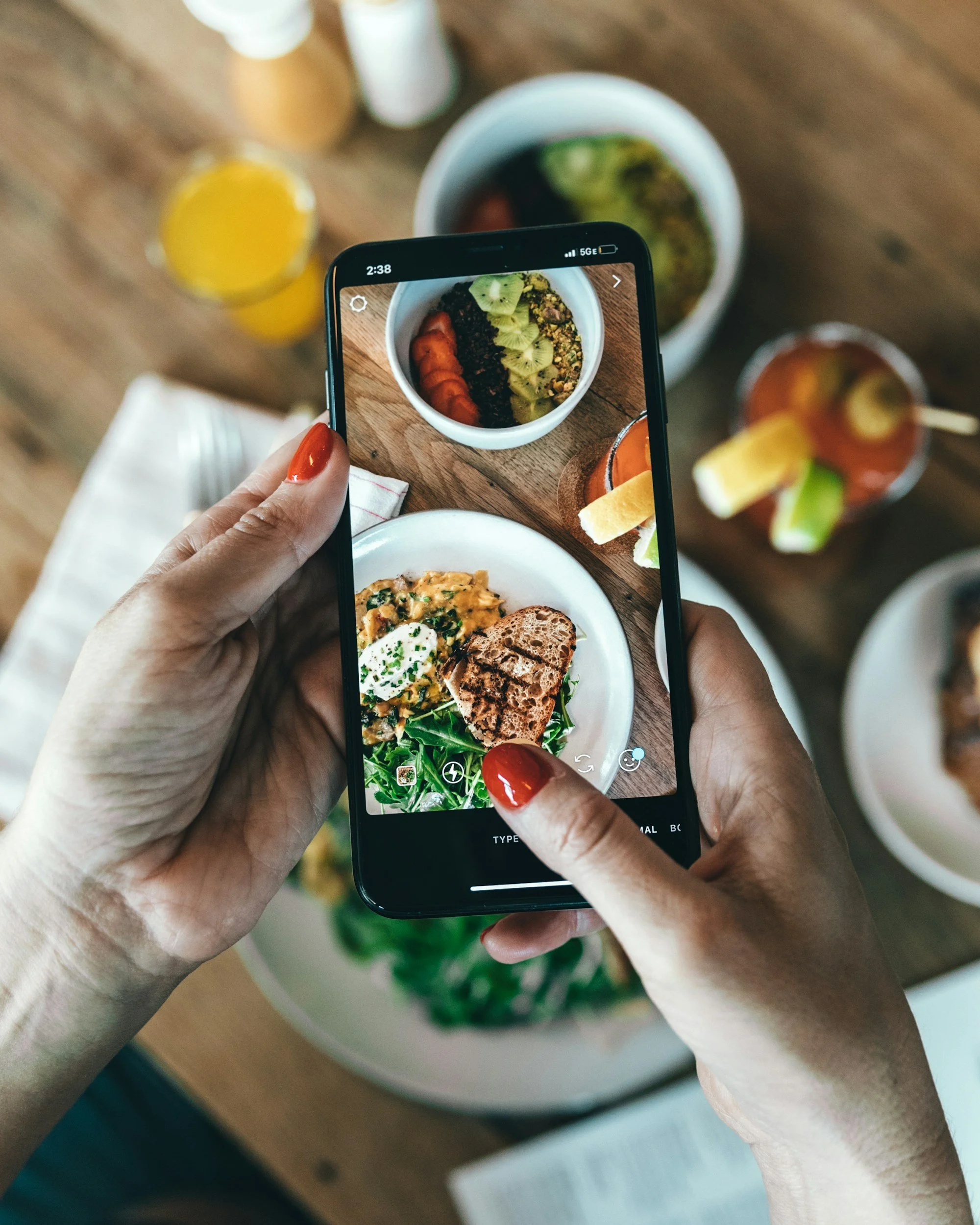 Person using smartphone to photograph a bowl of salad with sliced kiwi, tomatoes, and granola, with drinks and other dishes visible on the table.