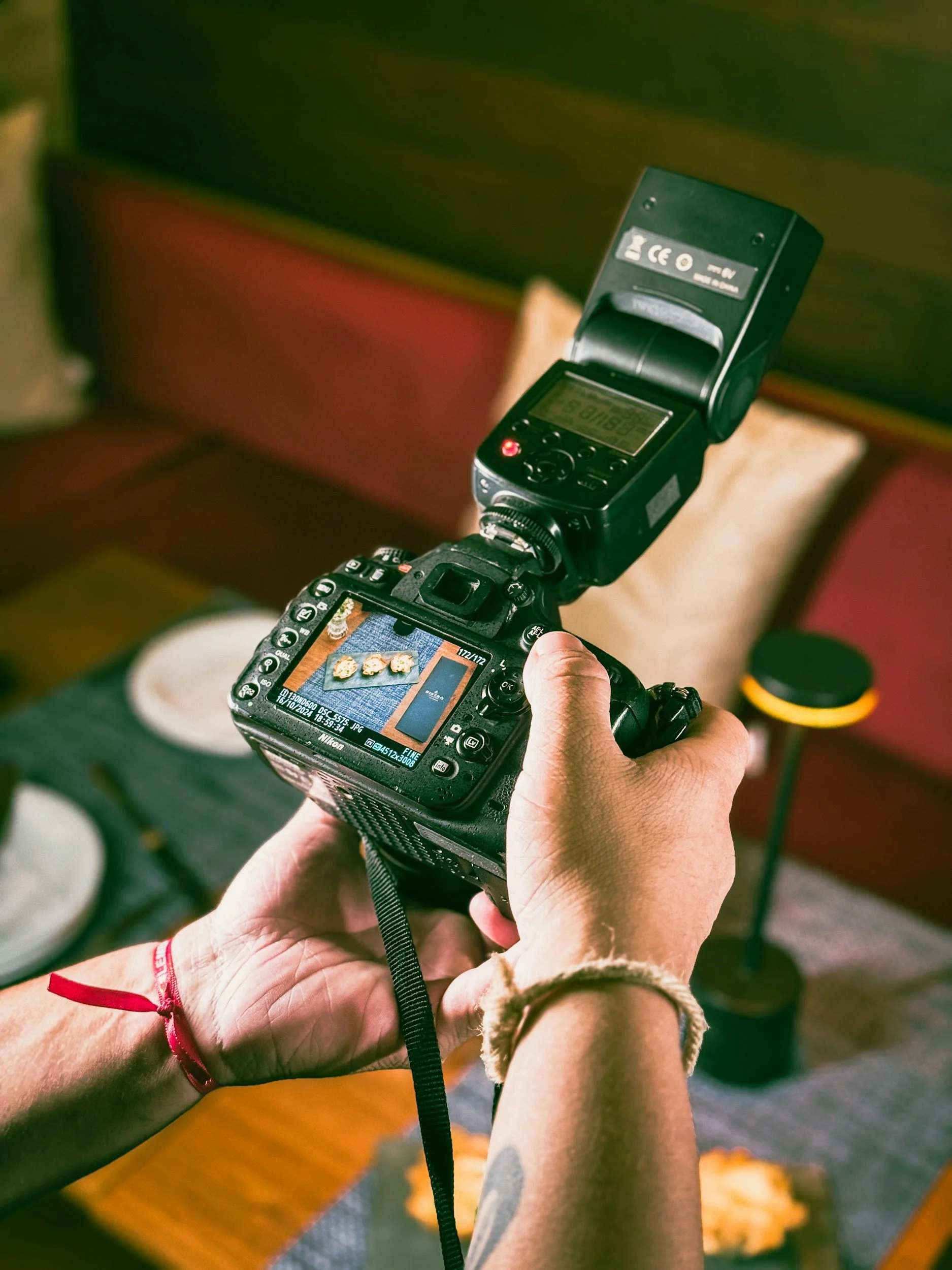 Close-up of a person holding a camera taking a photo of a dining table with food, with a handheld flash or light attached to the camera, and a person's forearm visible in the foreground.