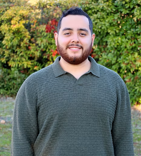 A young man with dark hair and a beard smiling outdoors in front of a colorful autumn foliage background.