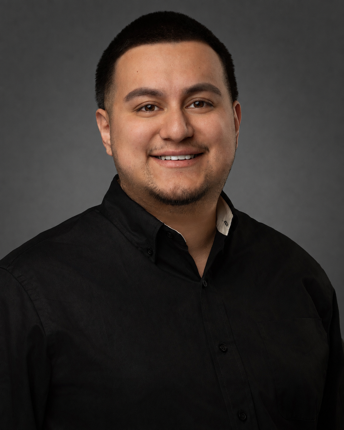 Headshot of a man with short dark hair, light skin, smiling, wearing a black collared shirt against a dark gray background.