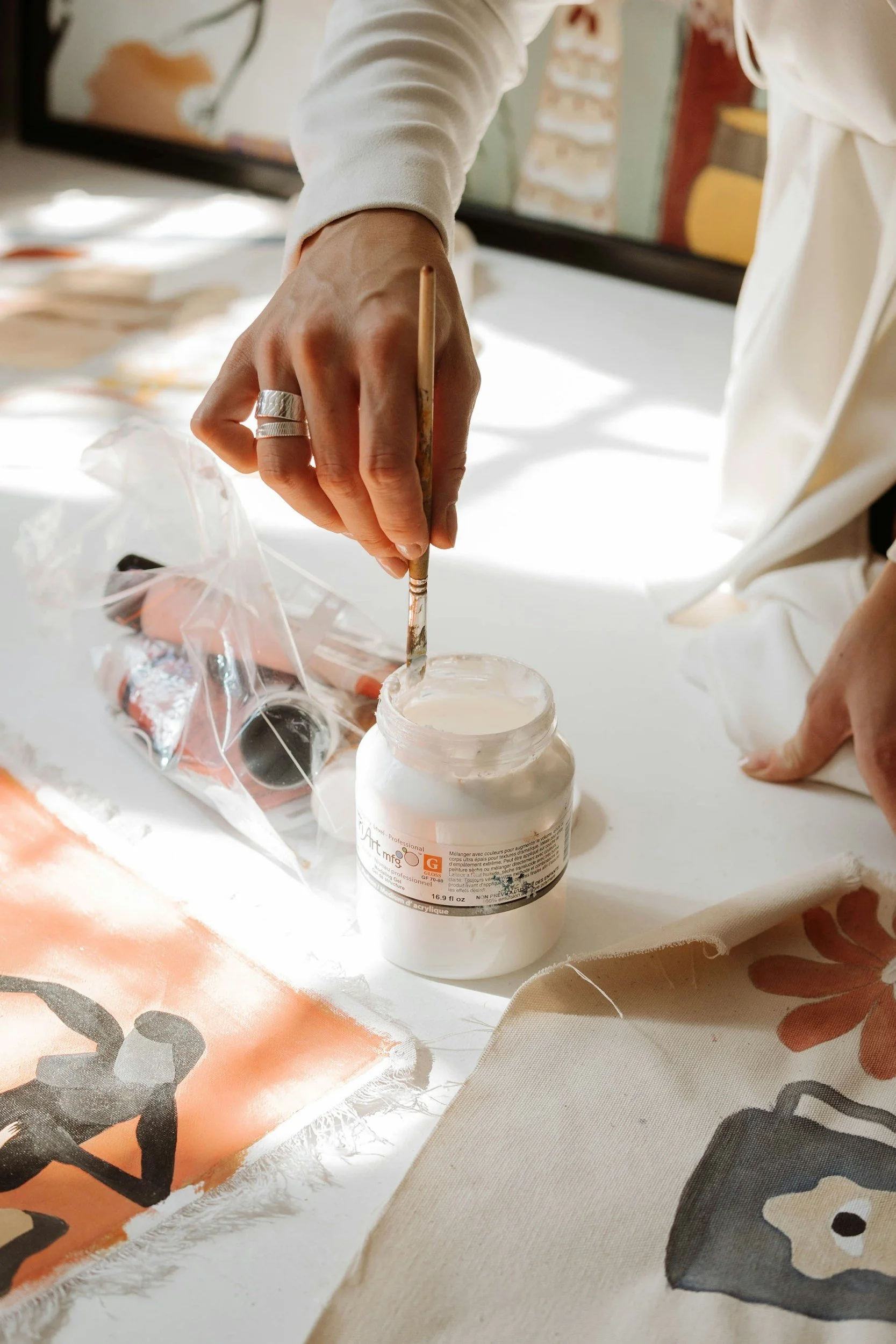 Close-up of a person using a paintbrush to apply white paint to a canvas. The person is wearing a white long-sleeve shirt and has a silver ring on their finger. The background shows a blurred image of a screen and some colorful artwork.