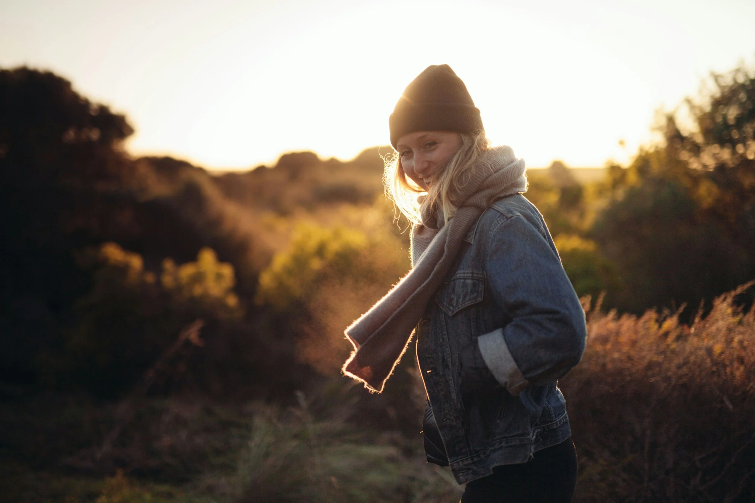 A young woman wearing a brown beanie, scarf, and denim jacket smiling outdoors during sunset.