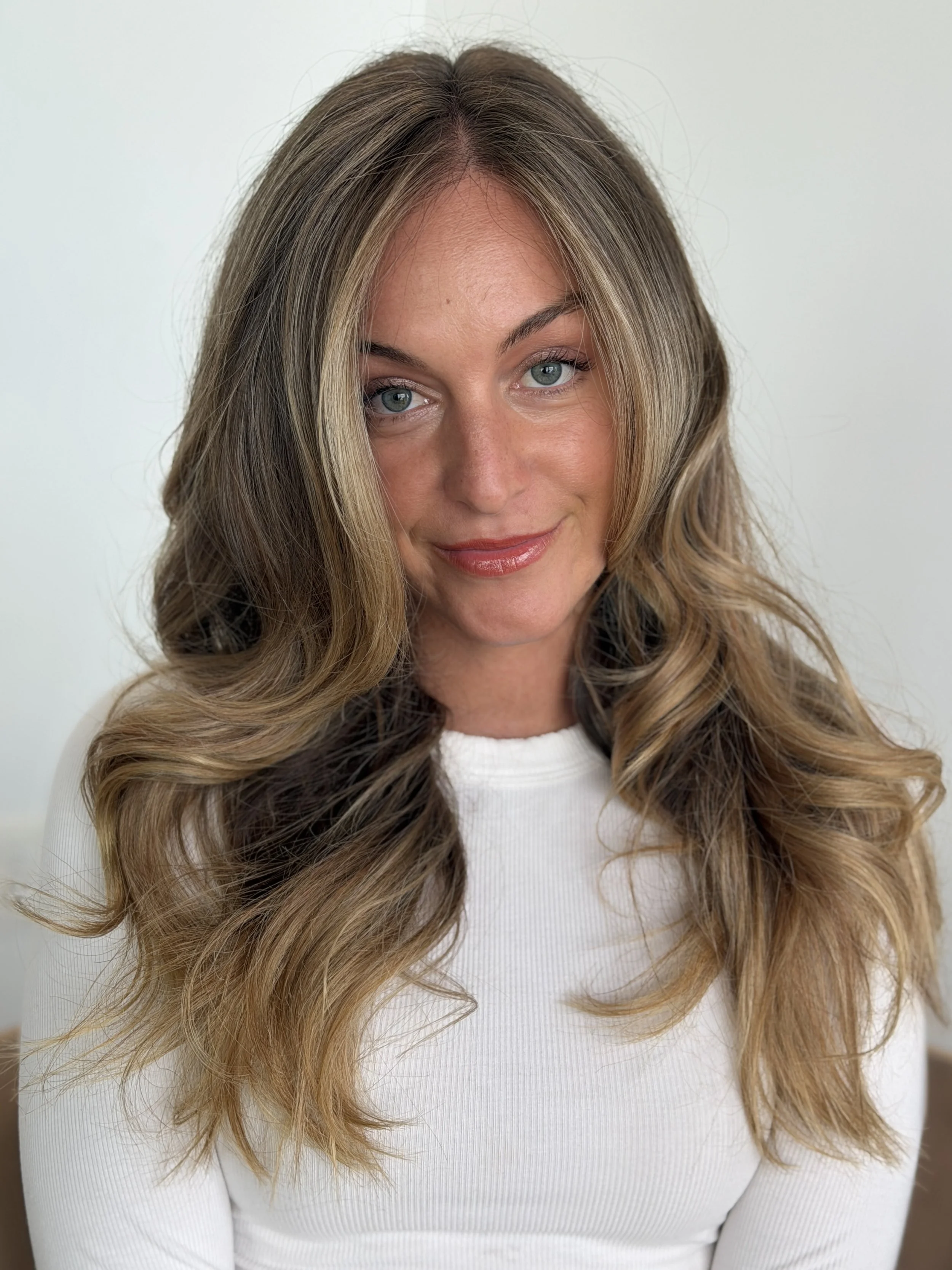 Close-up of a woman with long, wavy hair, blue eyes, and wearing a white top, smiling softly against a plain light background.