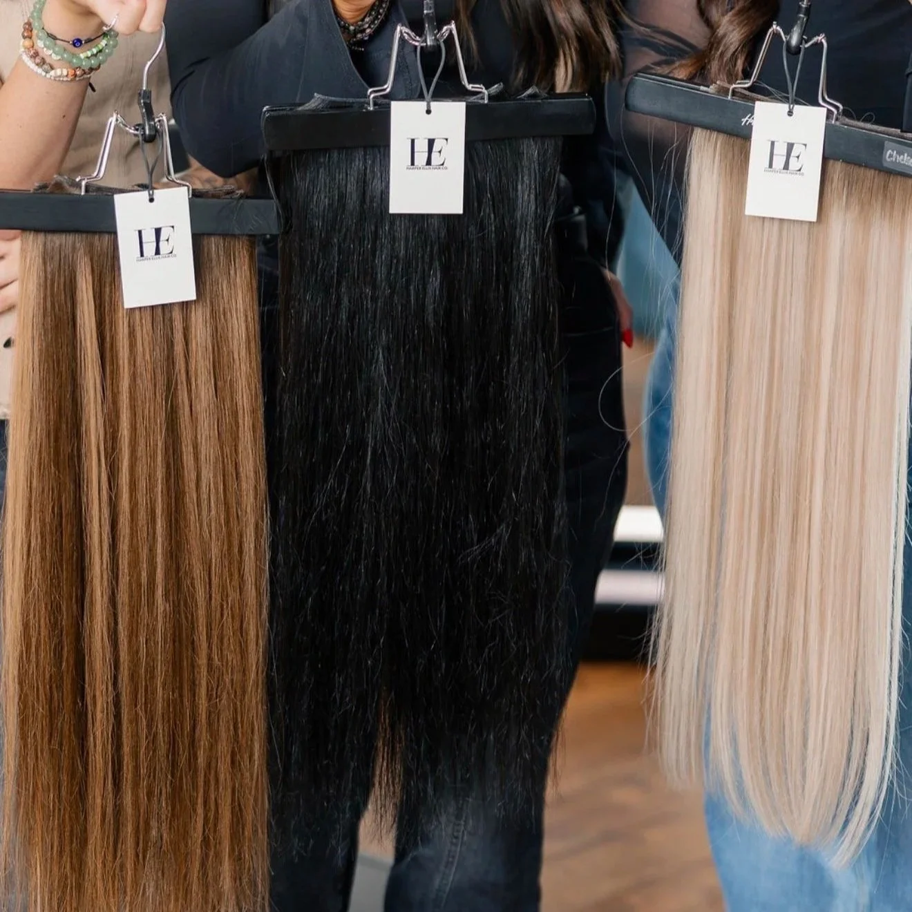 Harper Ellis Hair Co hair extensions swatches in different shades of blonde, brown, and black hanging on display hooks at a hair salon.