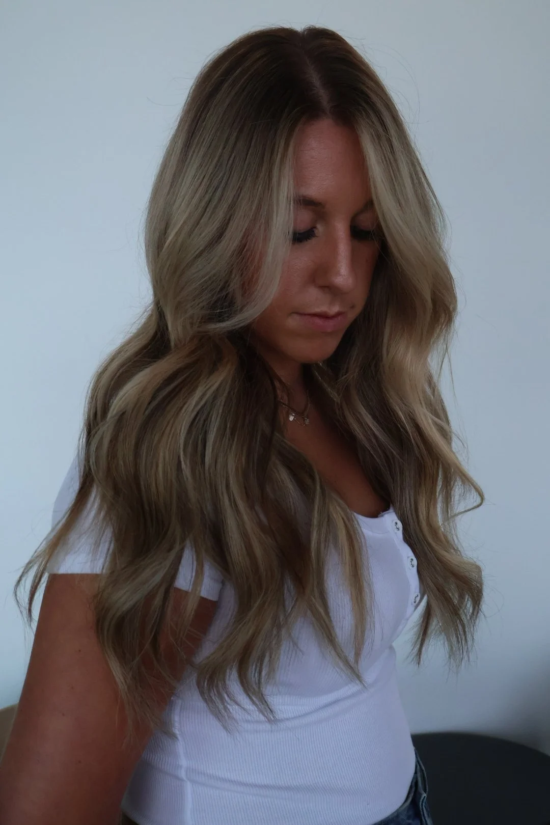 A woman with highlights, long, wavy blonde hair looking down, wearing a white shirt with buttons and a necklace, against a plain light-colored background.
