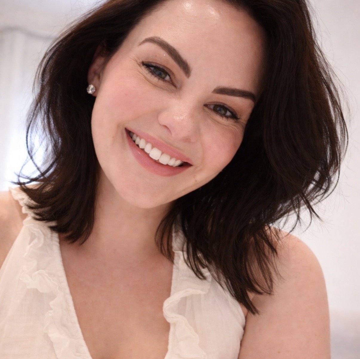 A woman with shoulder-length dark brown hair, smiling, wearing a sleeveless ruffled white top.