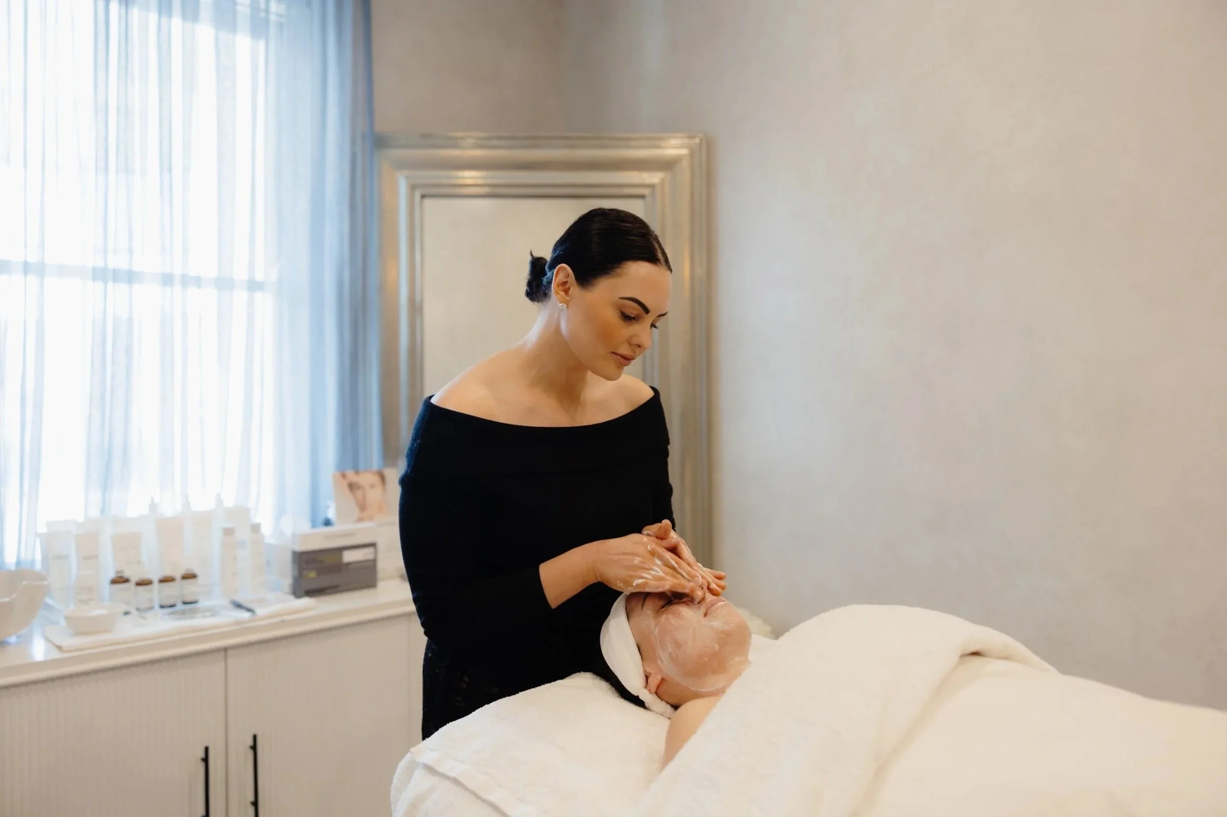A woman giving a facial treatment to a patient lying on a spa bed, covered with a white towel, in a bright, clean spa room with a window, curtains, and skincare products on a shelf.