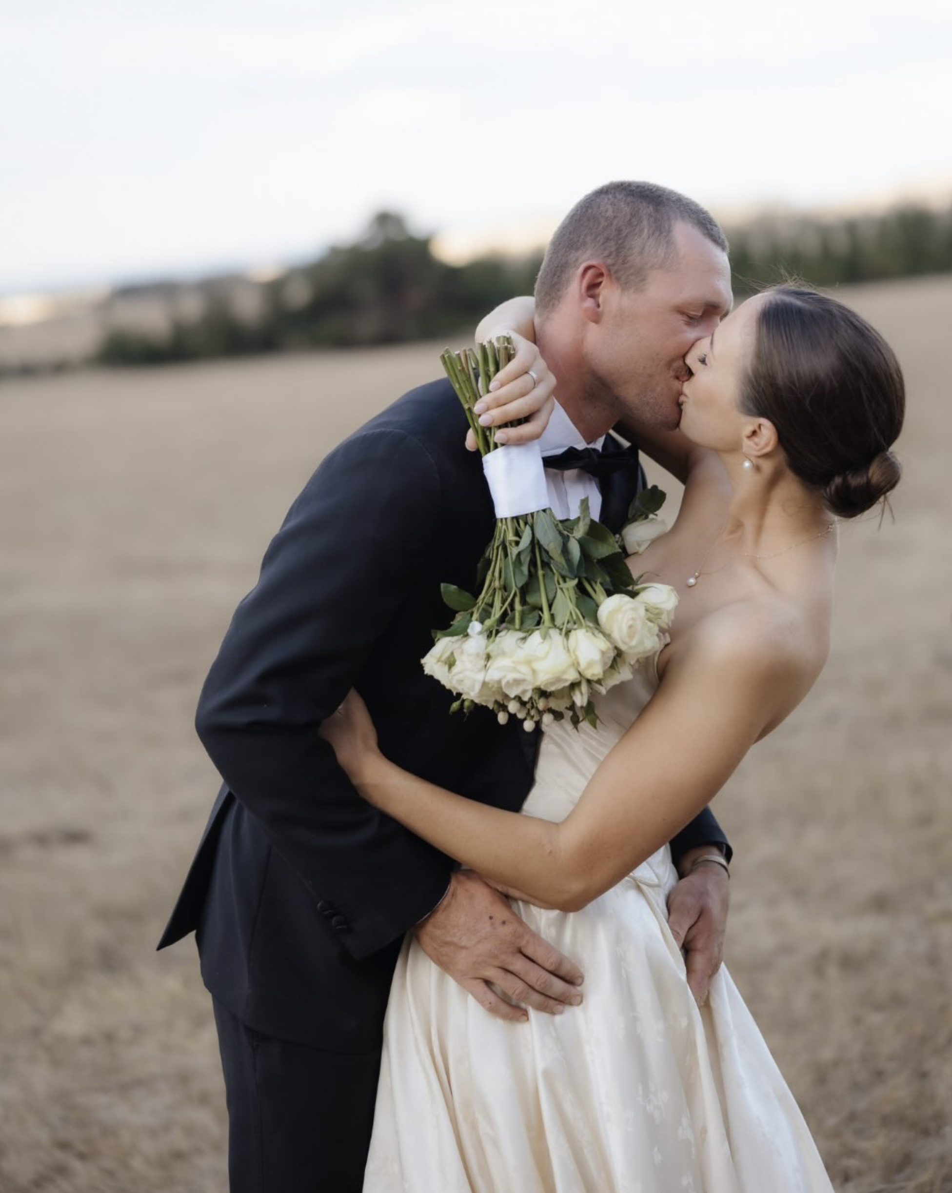 A newlywed couple sharing a kiss outdoors, with the groom in a tuxedo holding the bride and the bride in a wedding dress holding a bouquet of white roses.