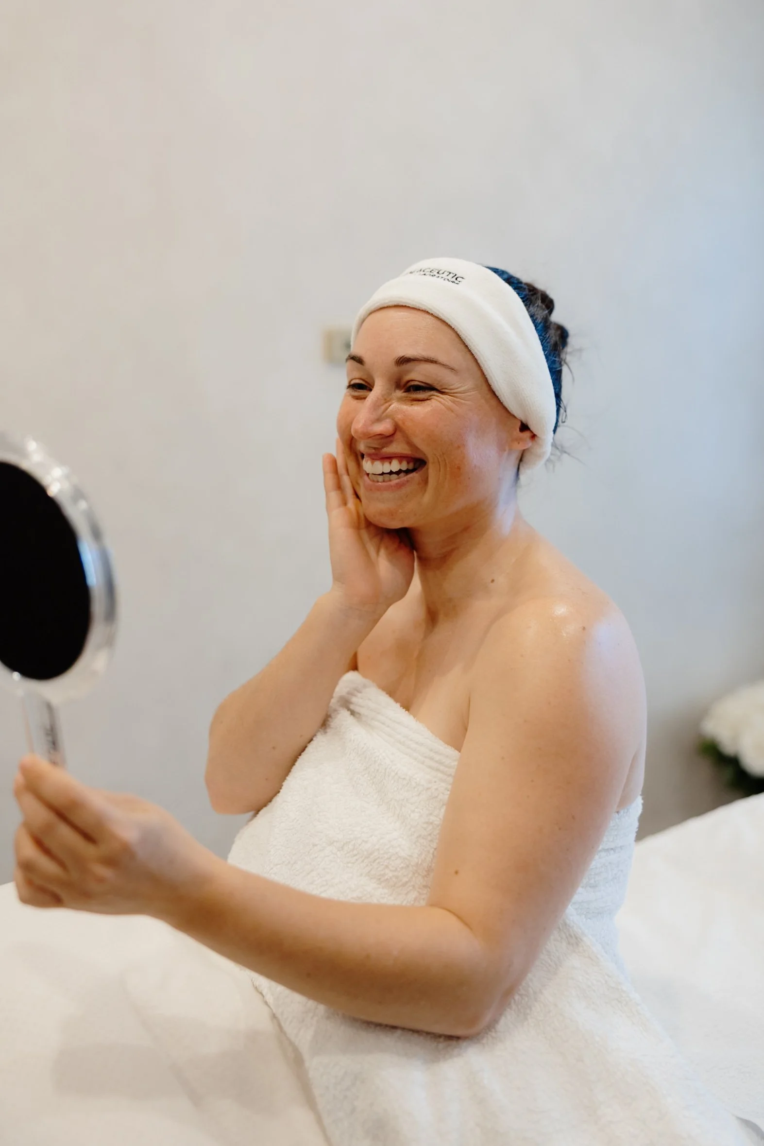 A woman wrapped in a white towel wearing a headband, smiling, looking at her reflection in a mirror, in a spa or bathroom setting.