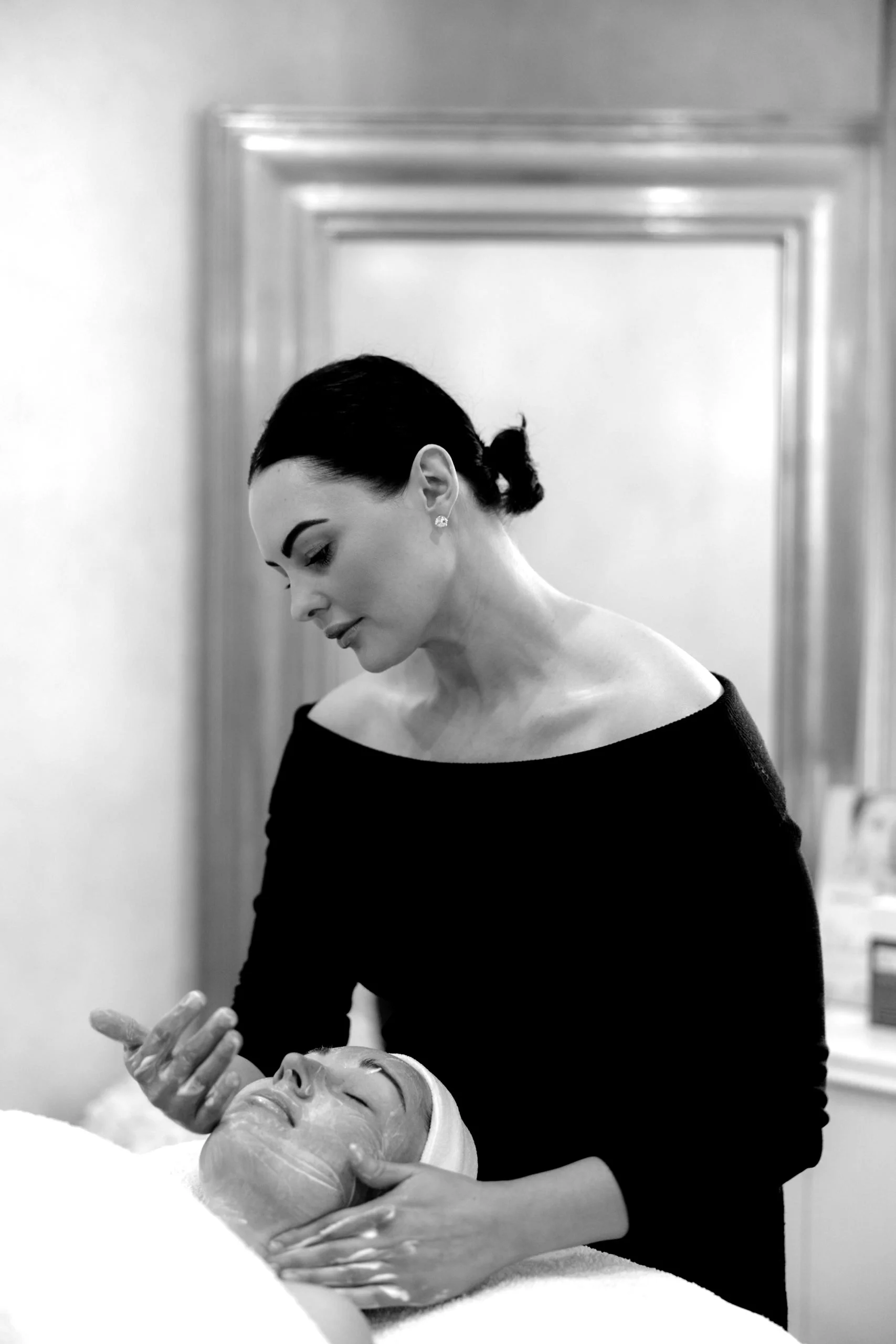 A woman giving a facial treatment to a woman lying down with her eyes closed, in a spa or beauty salon setting.