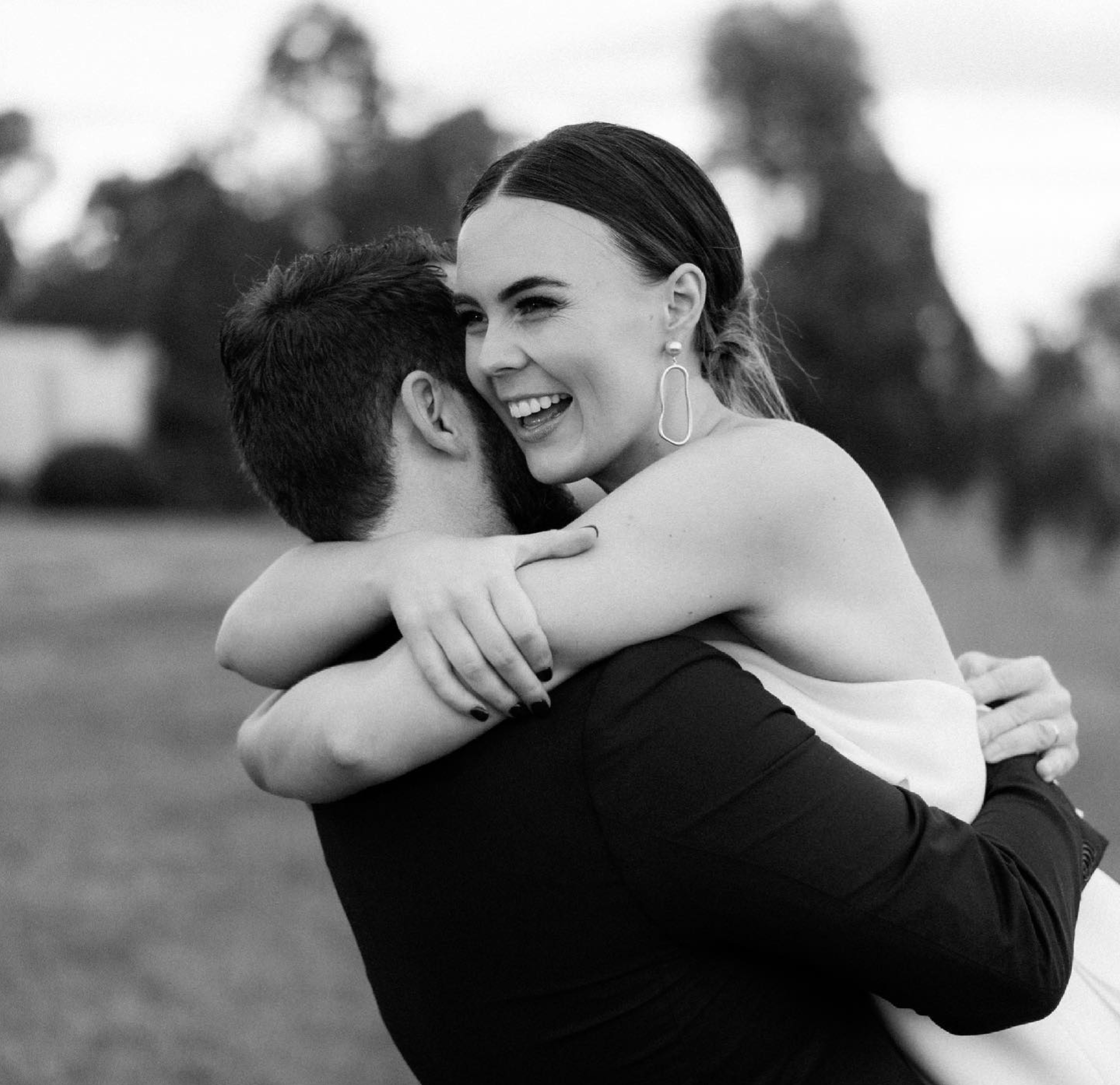 A black and white photograph of a couple embracing, with the woman smiling and wearing earrings, while the man is facing away from the camera.