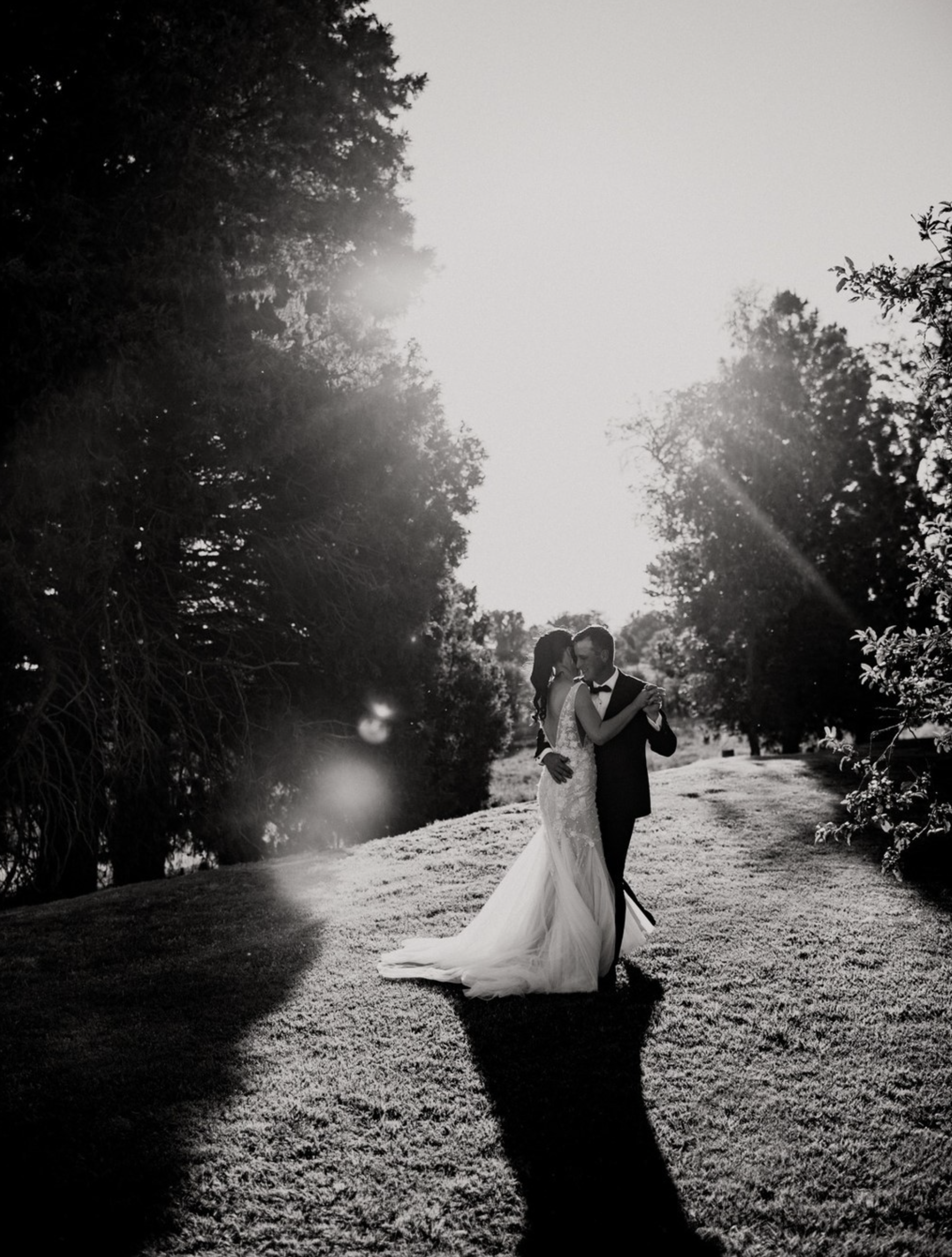 A black and white photo of a couple in wedding attire, embracing outdoors on a sunny day surrounded by trees and grass.