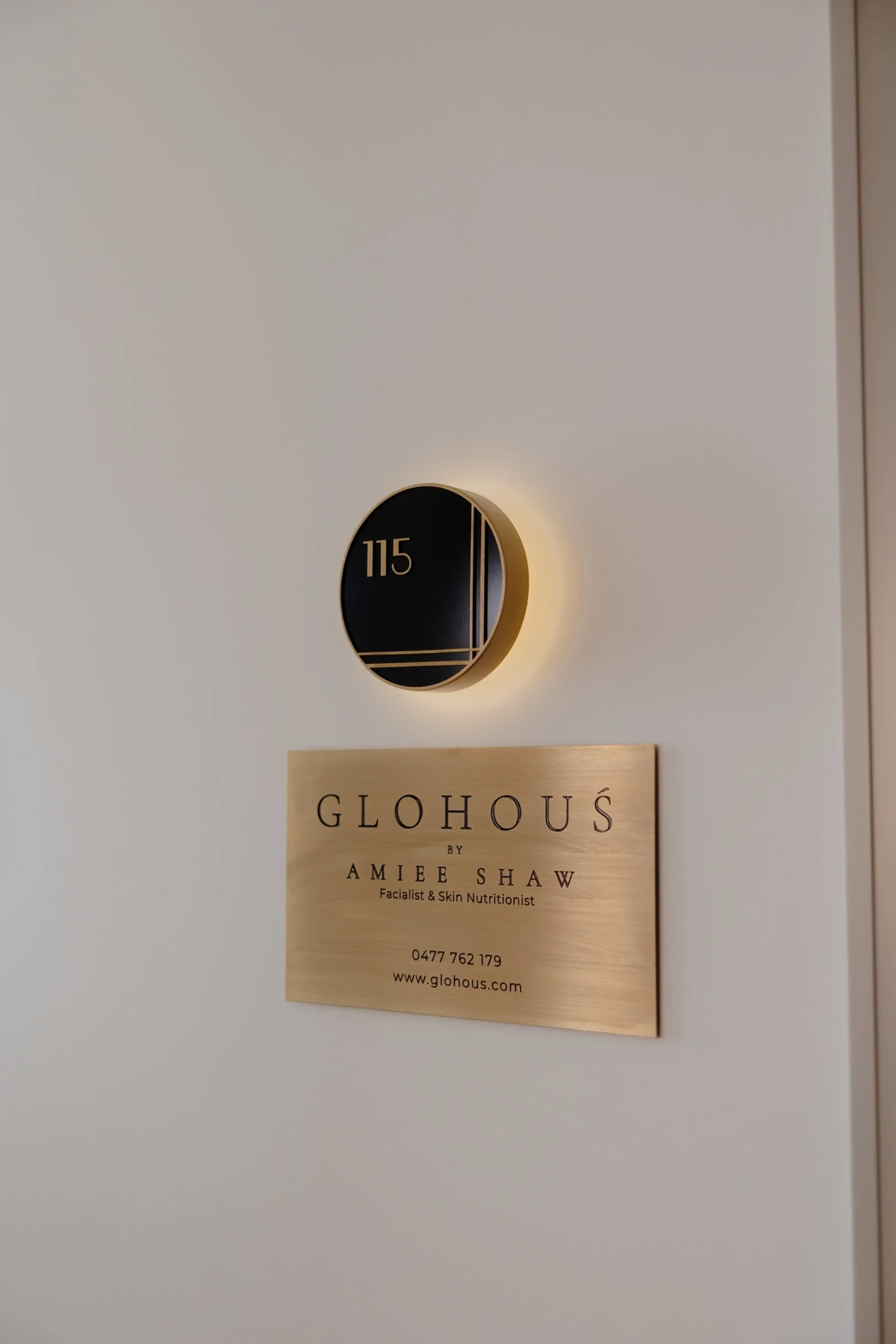 Wall-mounted black circular number 115 with backlit glow. Below is a gold-colored sign for GLOHOUS by Amiee Shaw, a facialist and skin nutritionist, with contact details and website.
