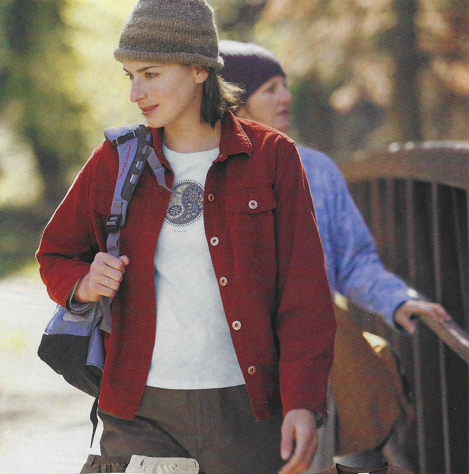 Two women walking outdoors with trees in the background. One woman in the foreground wears a gray beanie, red jacket, and white shirt, carrying a gray backpack. The other woman in the background wears a black beanie and a blue jacket.