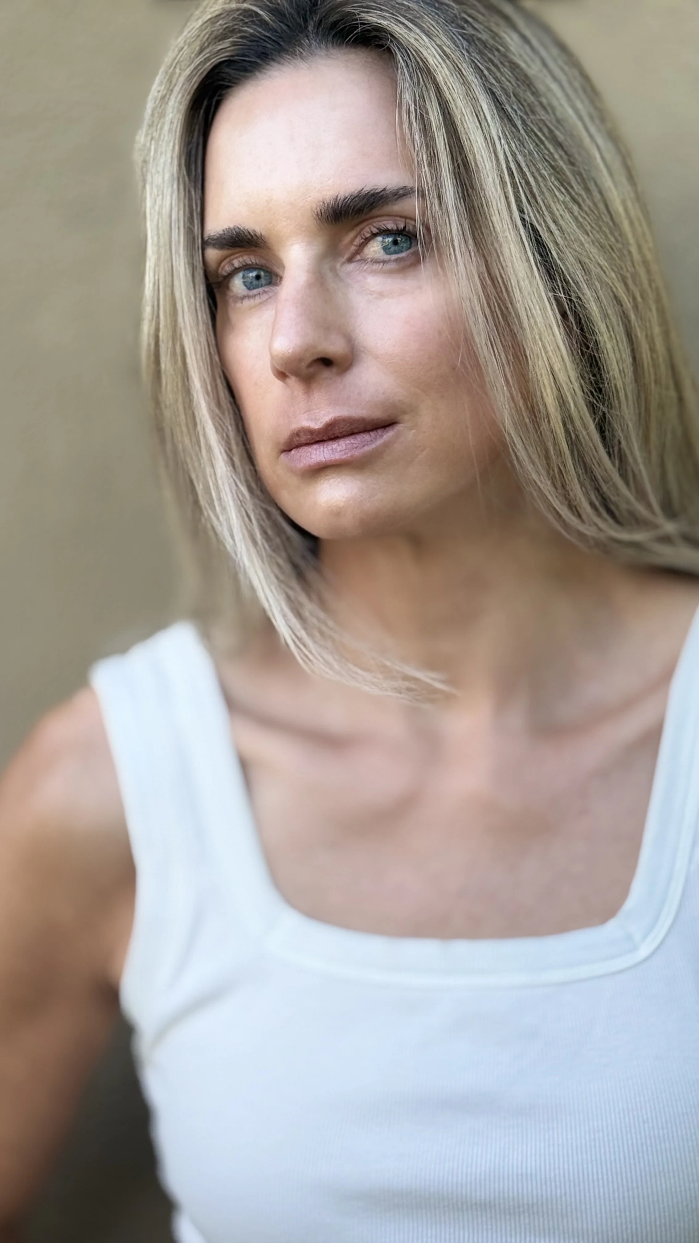 Close-up of a woman with blonde hair, blue eyes, and a light skin tone wearing a white sleeveless top.