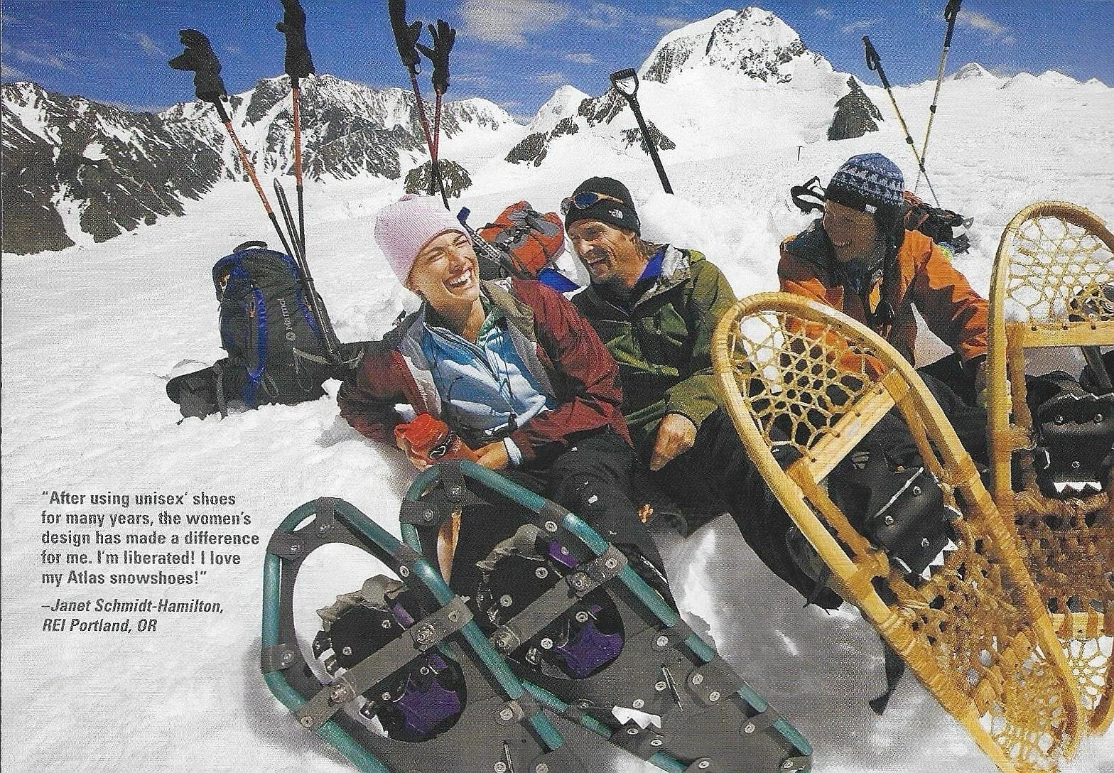 Three people sitting on snow in a snowy mountain landscape, smiling and laughing. Two are wearing winter jackets and hats, and one person has on snowshoes. Snowshoes are depicted in the foreground. There are backpacks behind them and mountain peaks in the background.