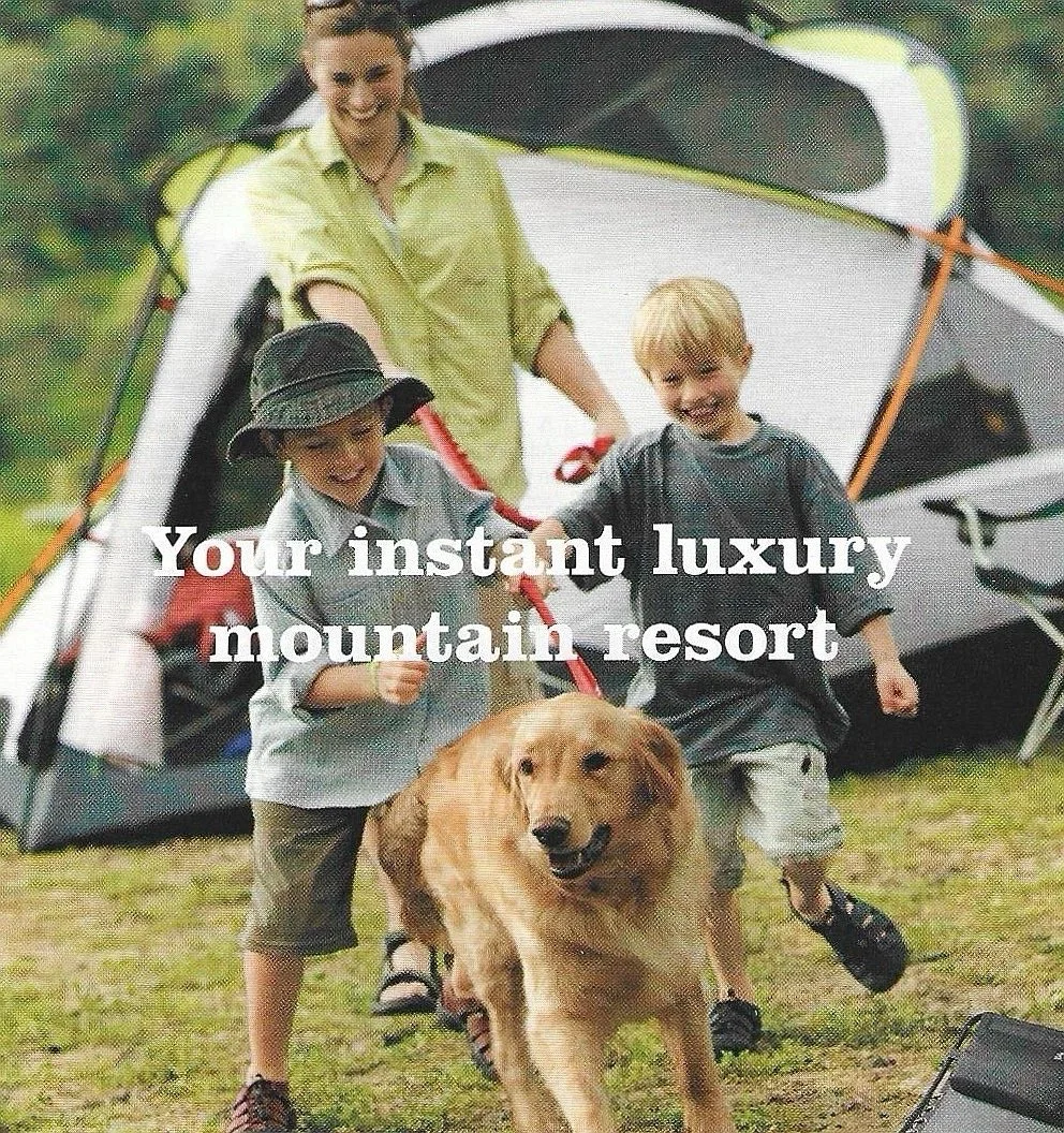 Two young boys and an adult woman with a dog at a camping site. The woman and boys are smiling and playing in front of a tent on a grassy area.