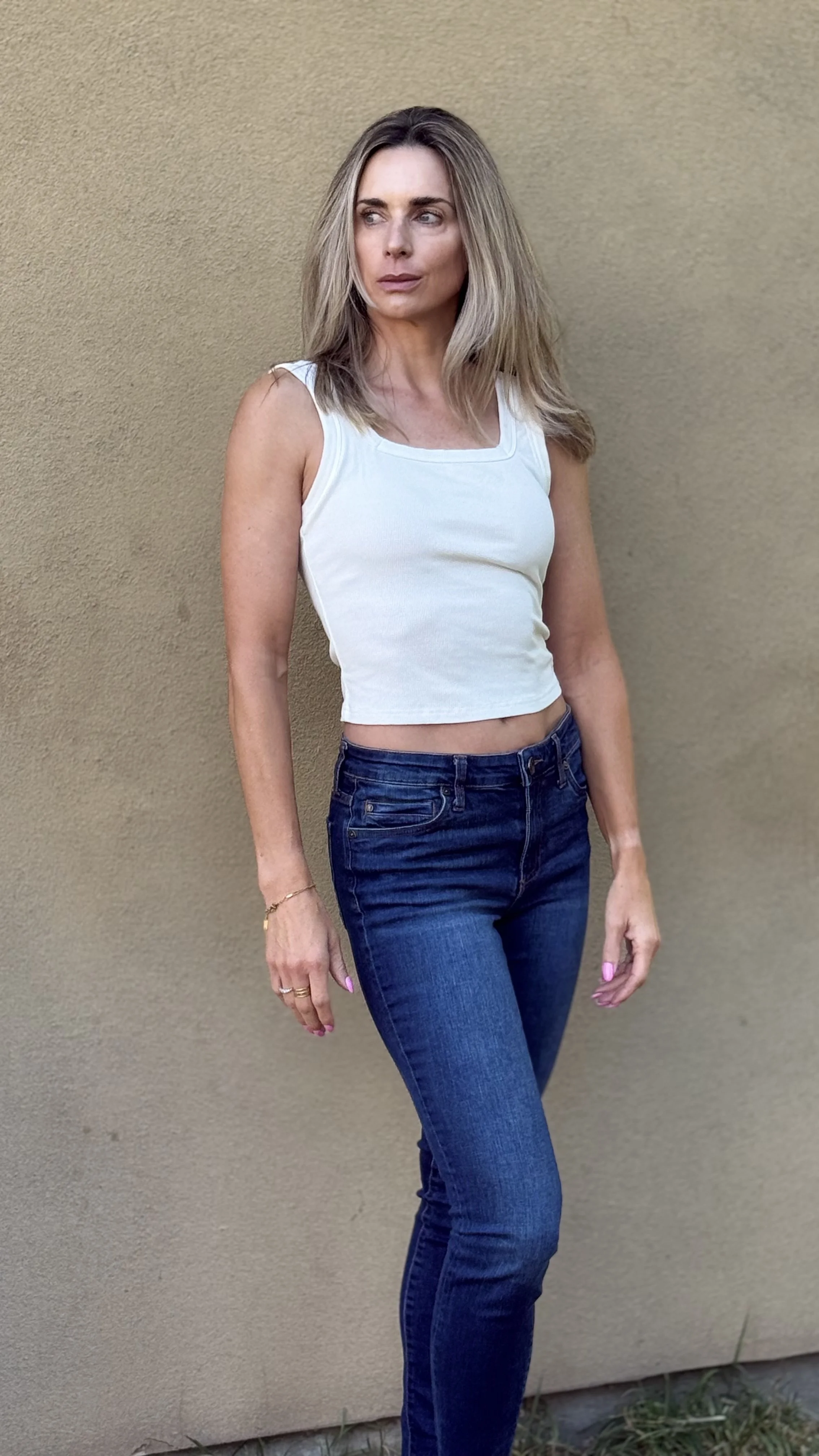 A woman with shoulder-length blond hair wearing a white sleeveless top and dark blue jeans standing against a beige wall.