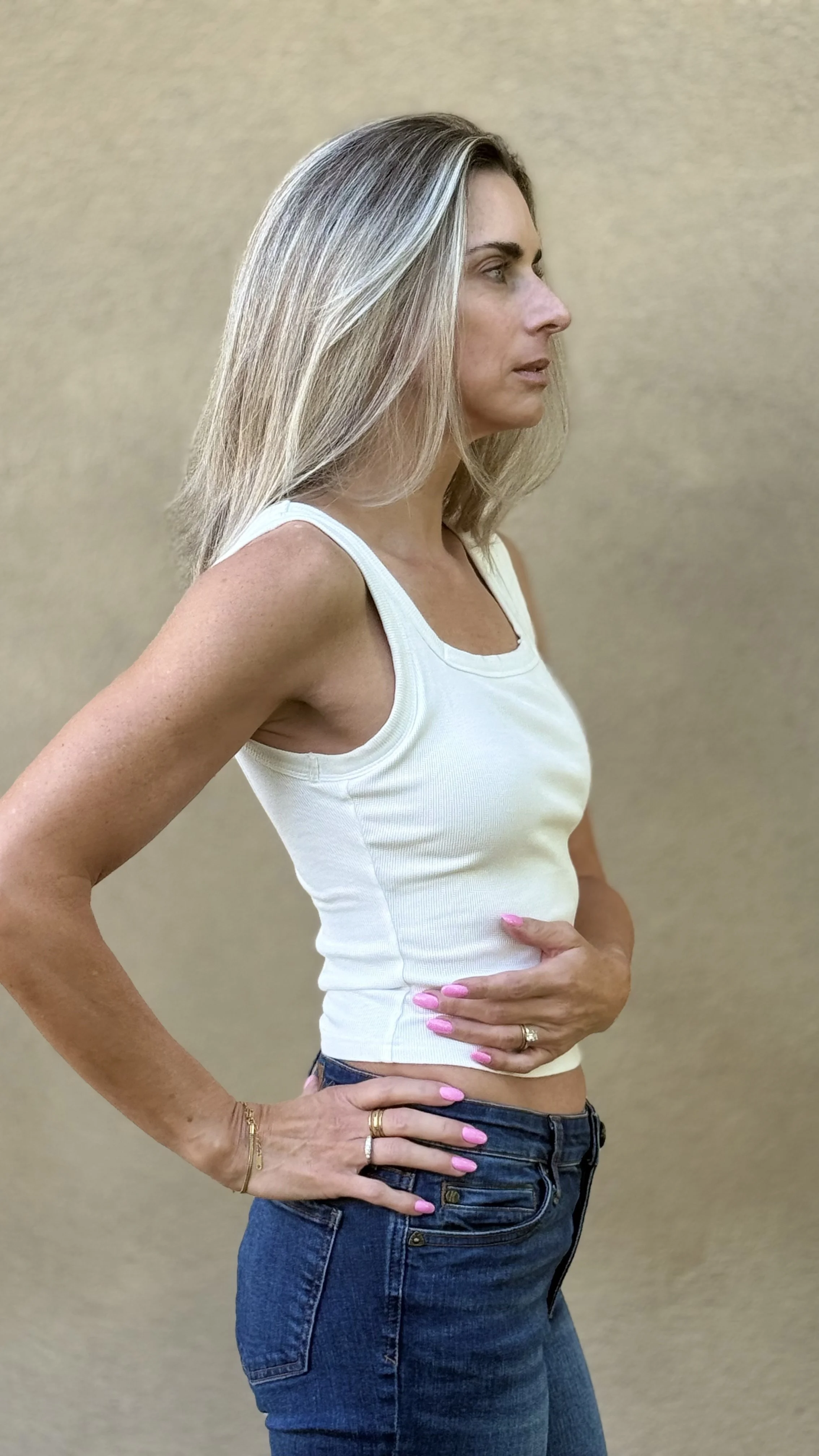 A woman with blonde hair in a white tank top and blue jeans stands sideways with her hands on her hips against a plain beige background.
