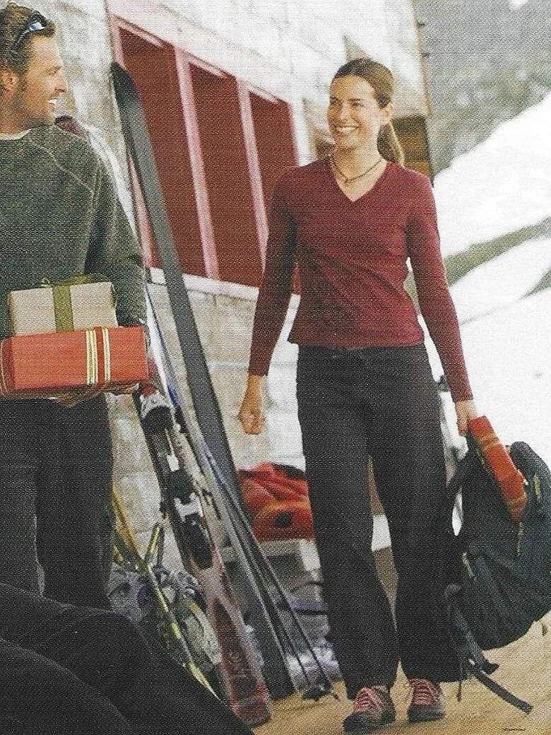 Two women standing outdoors near a wooden building, chatting and smiling, with snow visible in the background; one woman holds a bag and the other with a gift box.