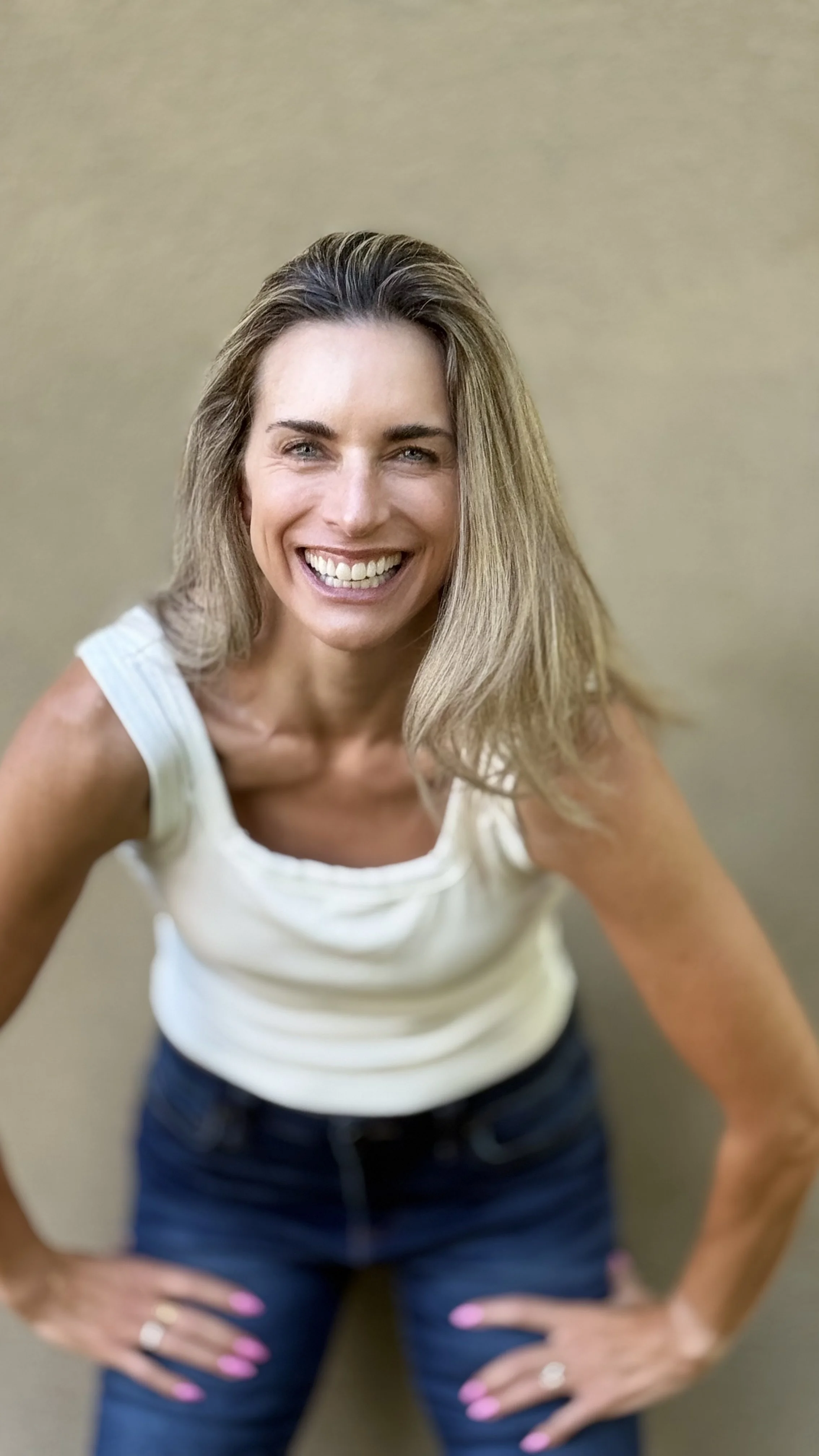 A woman with long blonde hair smiling, wearing a white sleeveless top and dark blue jeans, with her hands on her hips, standing against a neutral background.