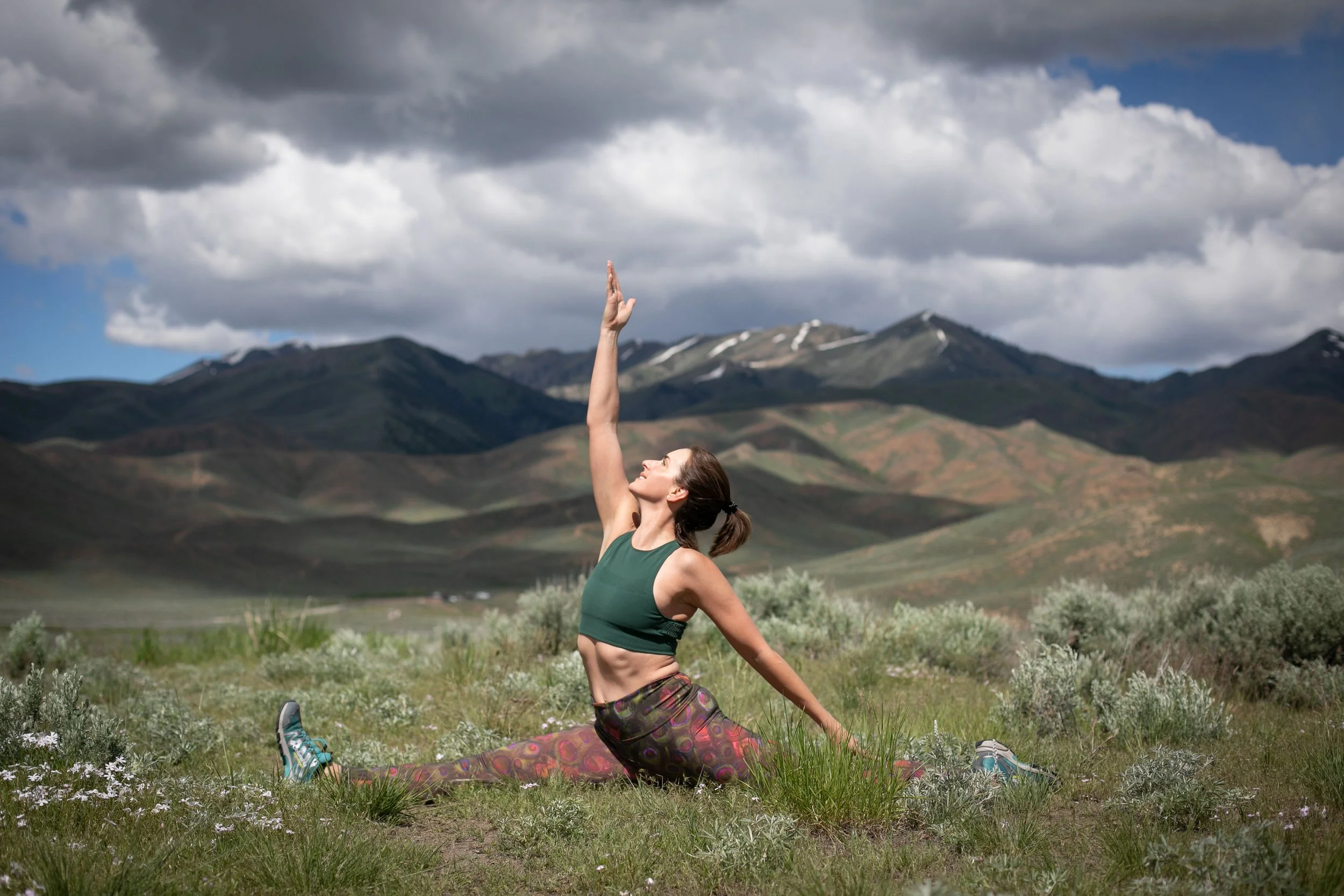A woman in colorful workout clothes performing a yoga pose on grass in an open field with mountains and a cloudy sky in the background.