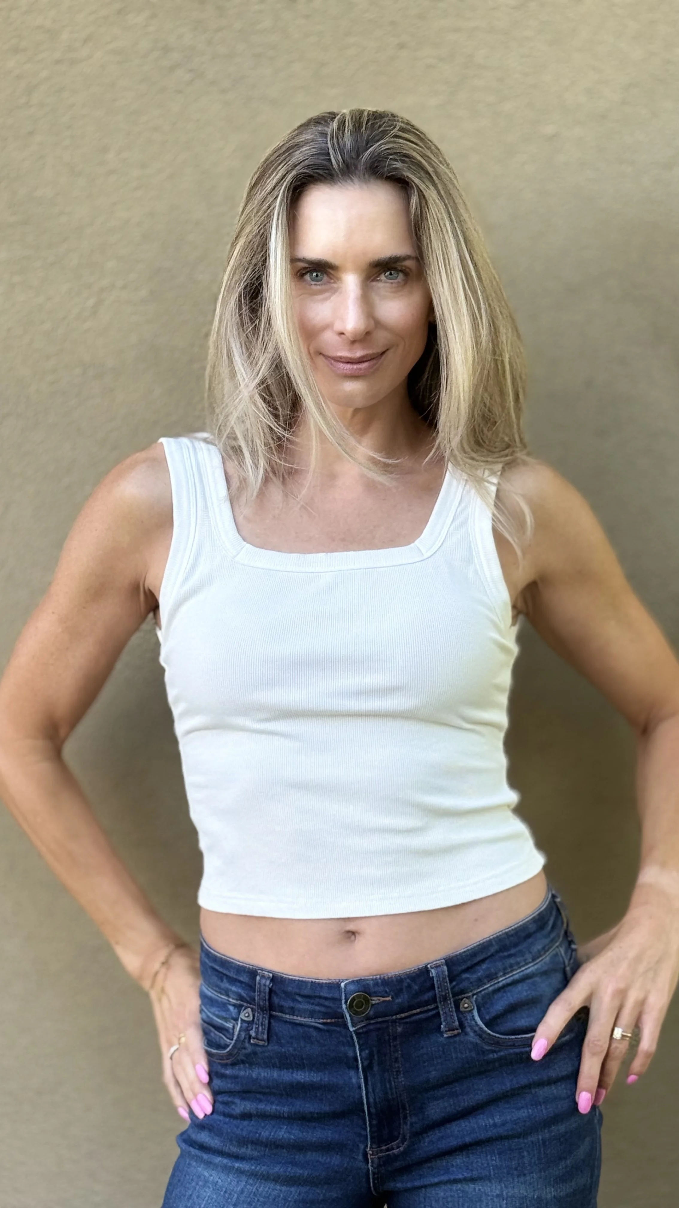 A woman with blonde hair and blue eyes, wearing a white sleeveless top and blue jeans, standing with hands on hips against a plain beige wall.