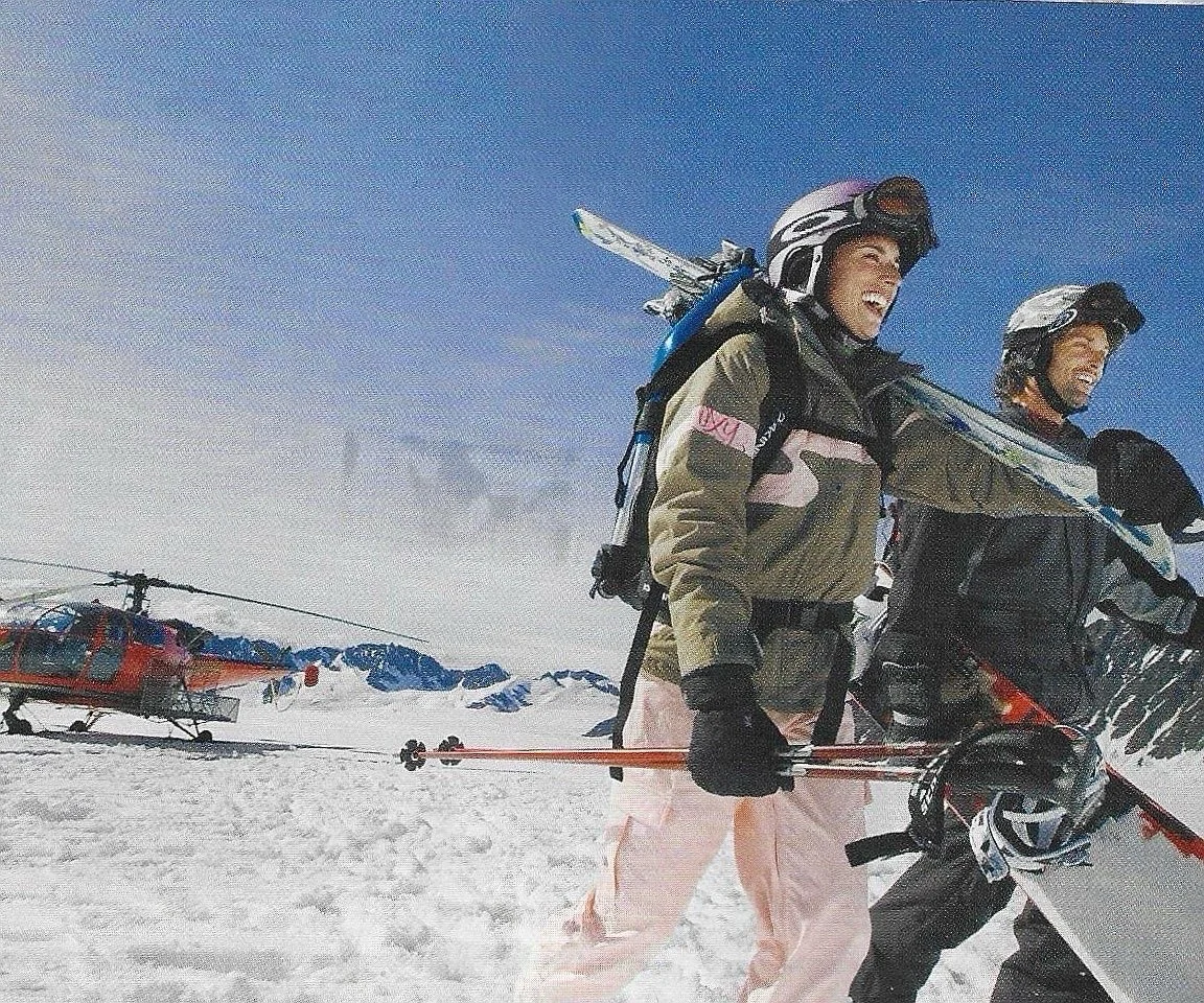 Two skiers in winter gear smiling with a helicopter in the snowy mountains behind them