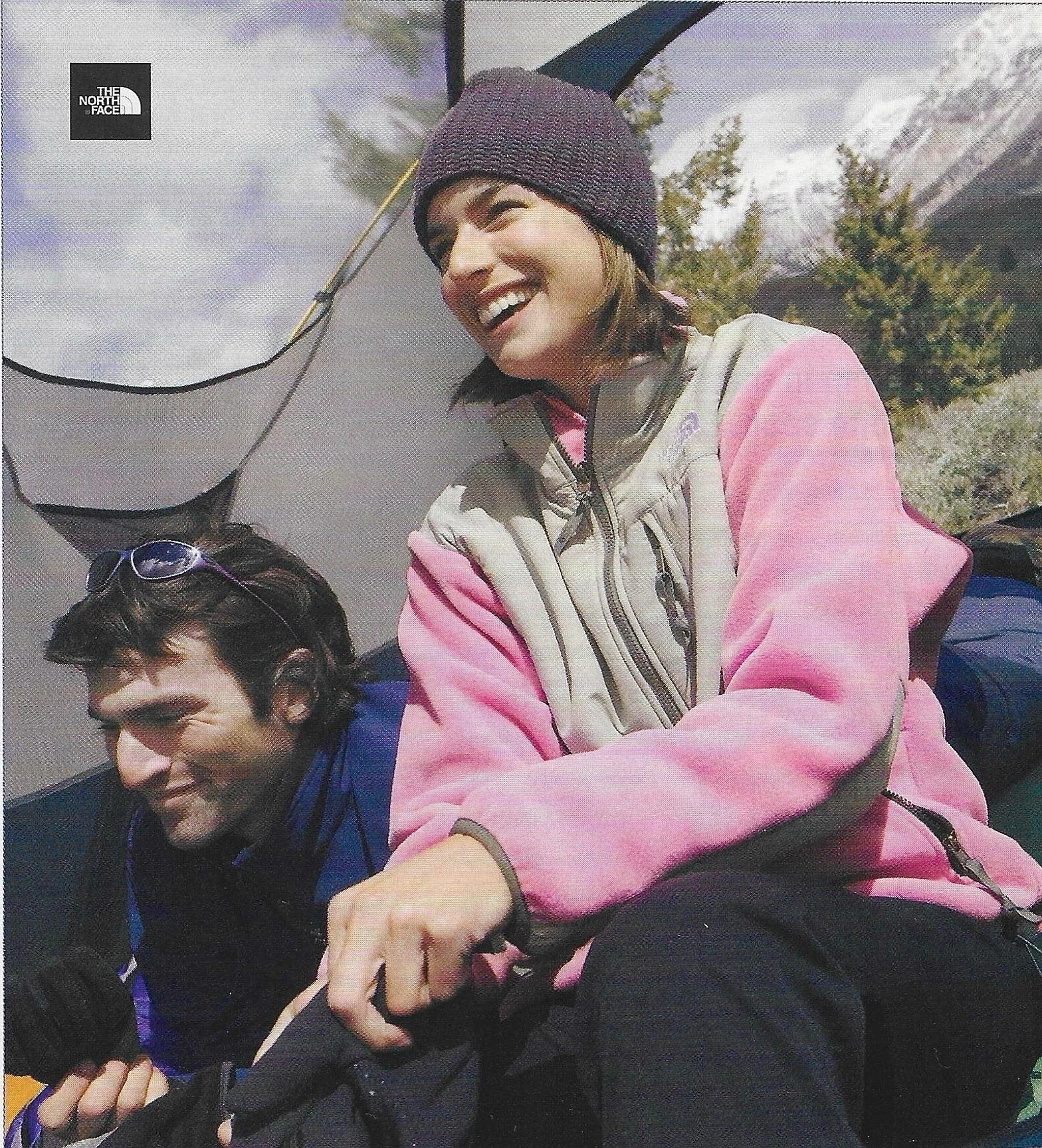 A woman wearing a purple beanie and a pink and beige jacket, smiling and laughing while riding on the back of a man with glasses and dark hair, with a mountainous outdoor background.