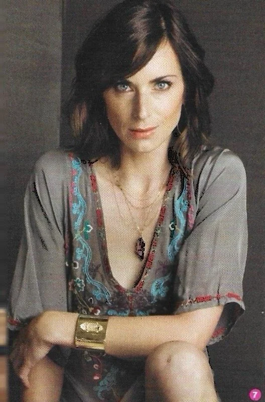 A woman with shoulder-length dark brown hair and blue eyes, wearing a gray top with colorful embroidery, a gold bracelet, and a necklace with a purple pendant, sitting with her arms crossed on her knee.