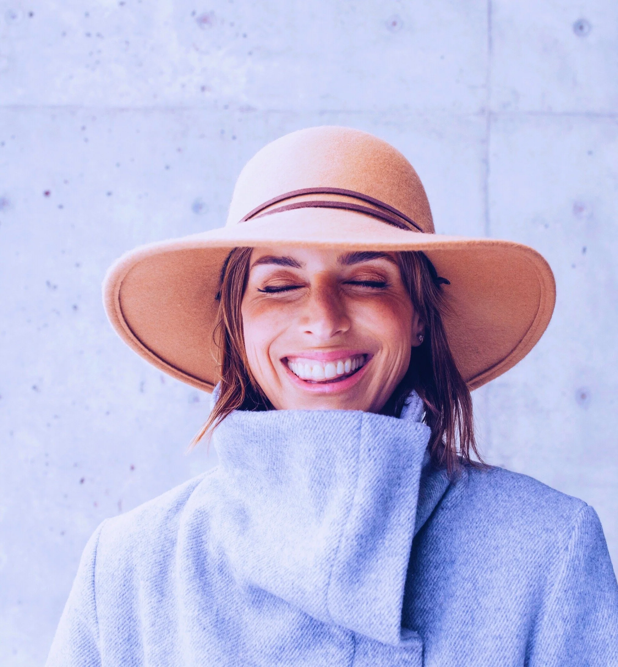 A woman smiling with eyes closed, wearing a wide-brimmed tan hat and a light blue sweater against a light-colored wall background.