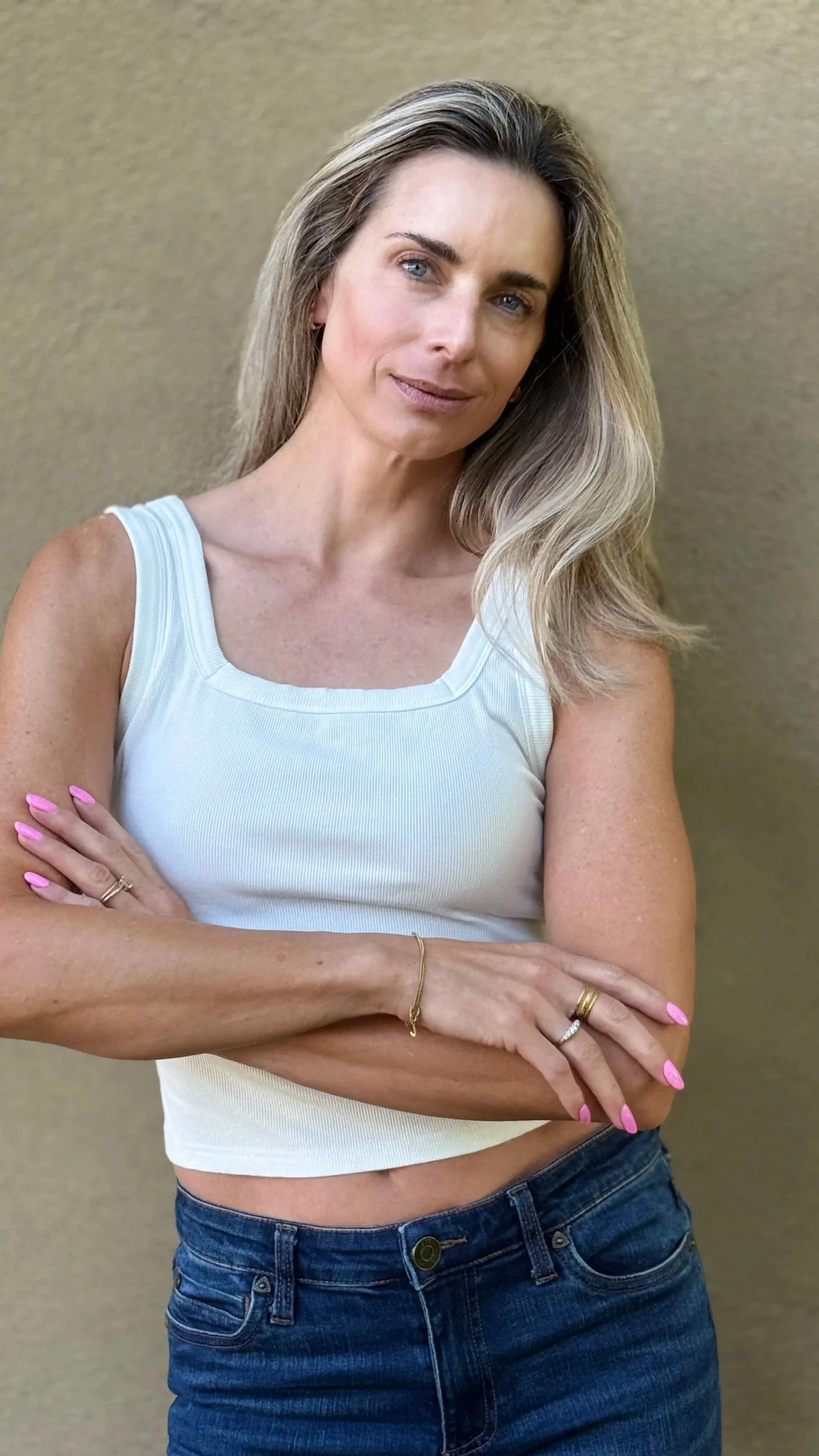A woman with long blond hair, blue eyes, wearing a white sleeveless top and dark jeans, standing with crossed arms against a beige background.