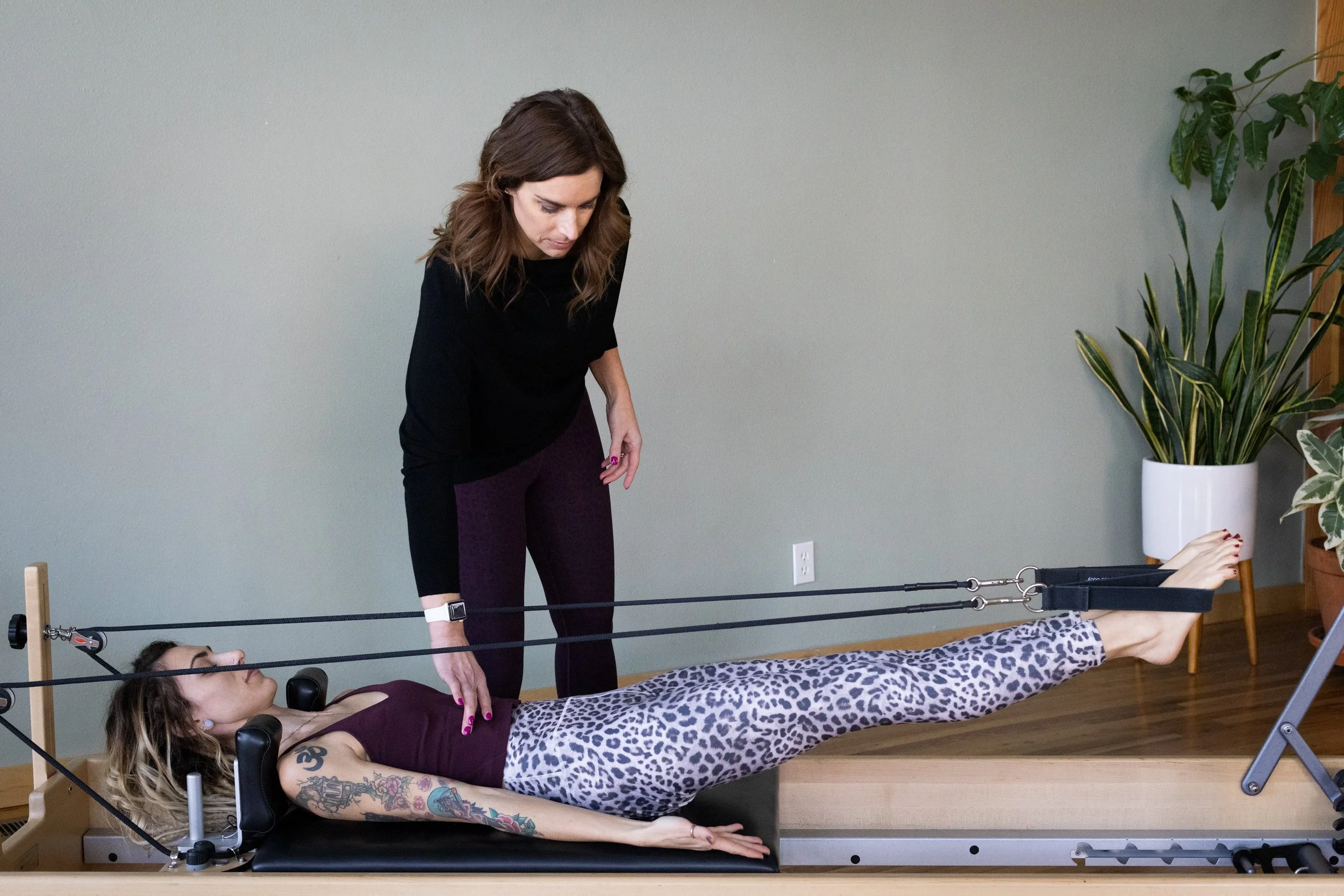 A woman with tattoos on her arm lying on a Pilates reformer machine, doing a leg stretch exercise, while a trainer stands beside her in a fitness studio.