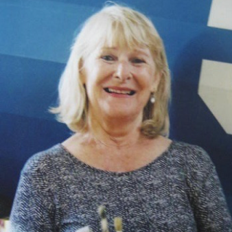 An elderly woman with blonde hair smiling, wearing a sparkly top, in front of a blue background.